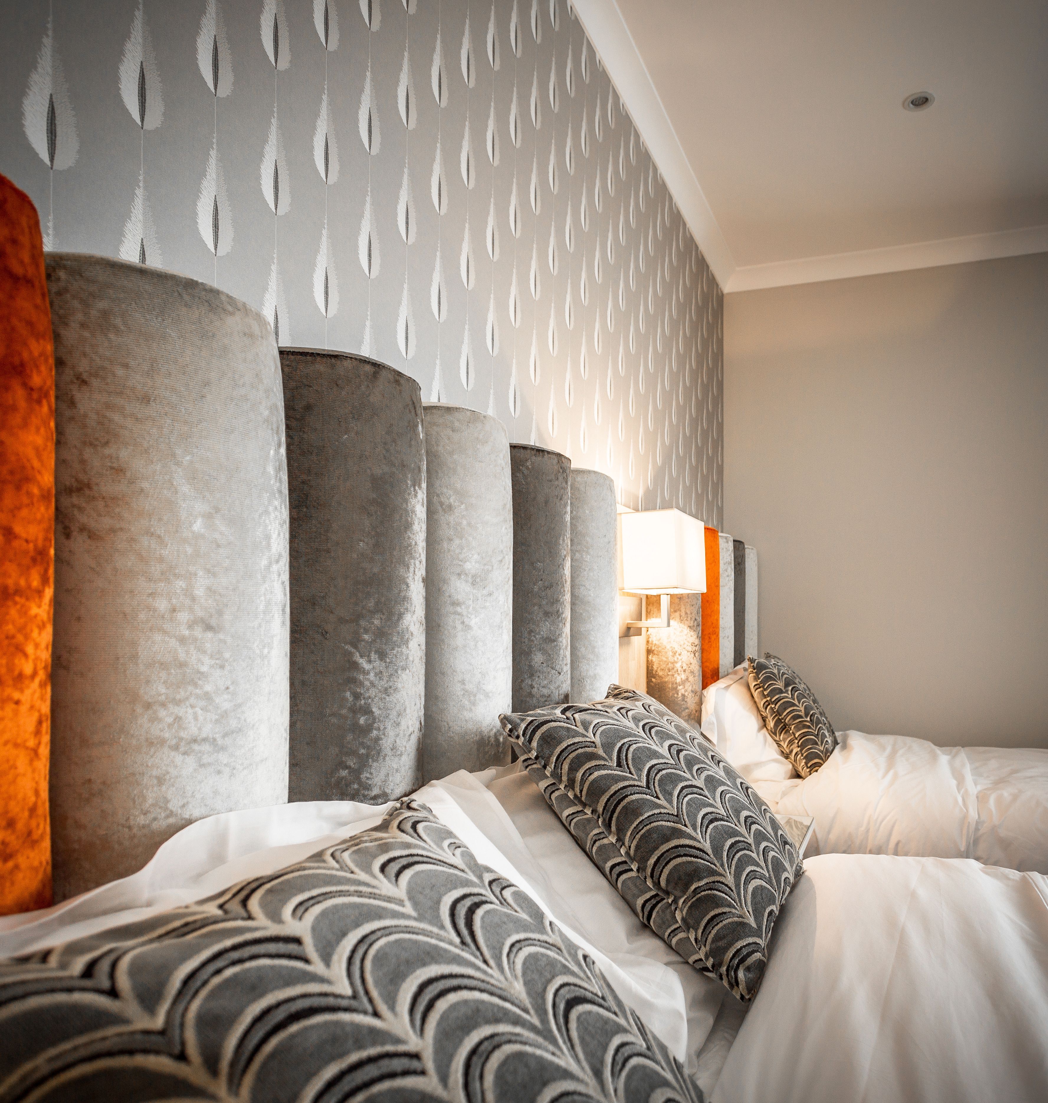 A modern hotel room with a large bed, multiple pillows, a nightstand with a lamp, and a patterned wallpaper.