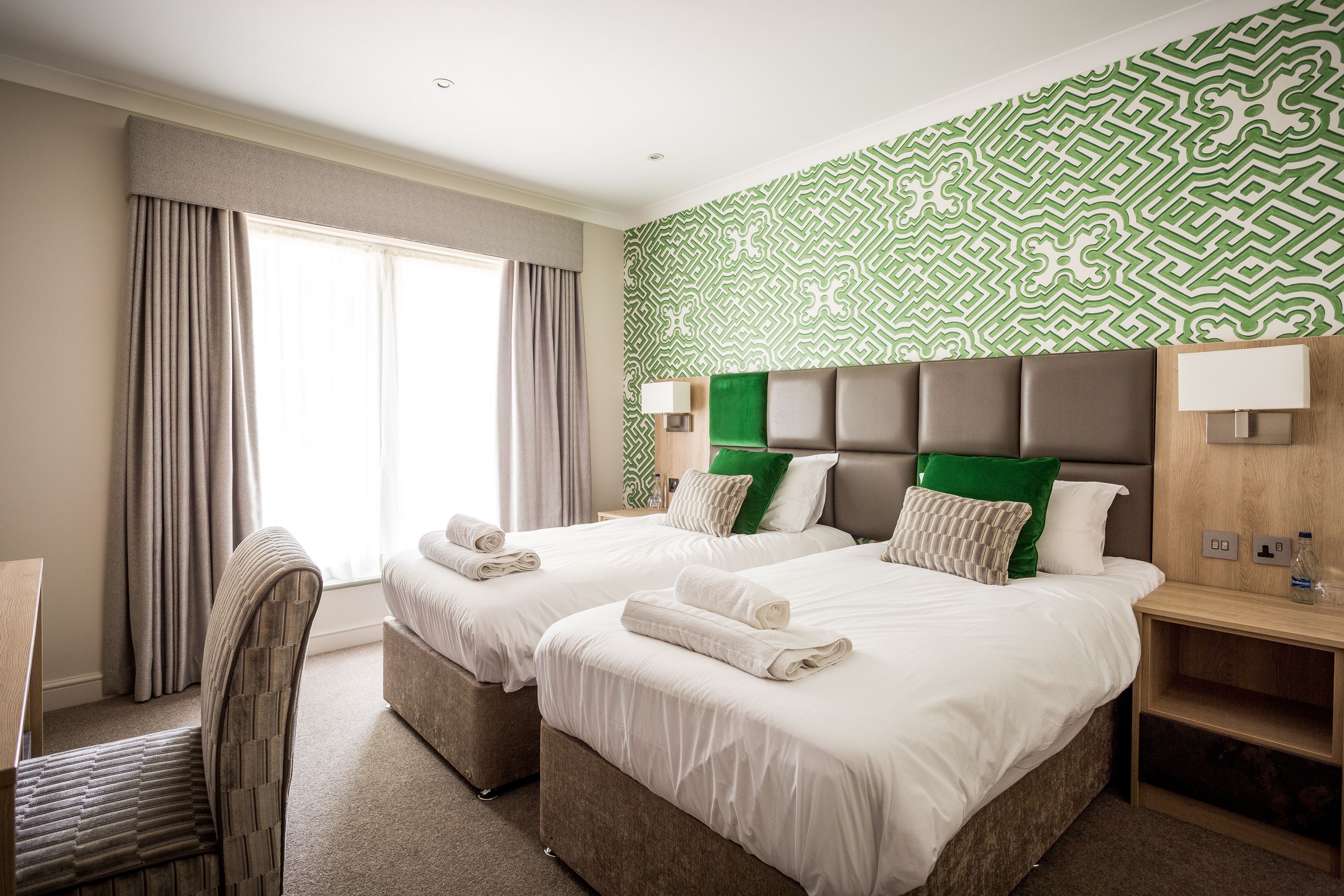 A hotel room with two beds, a desk, a chair, and a patterned wall.