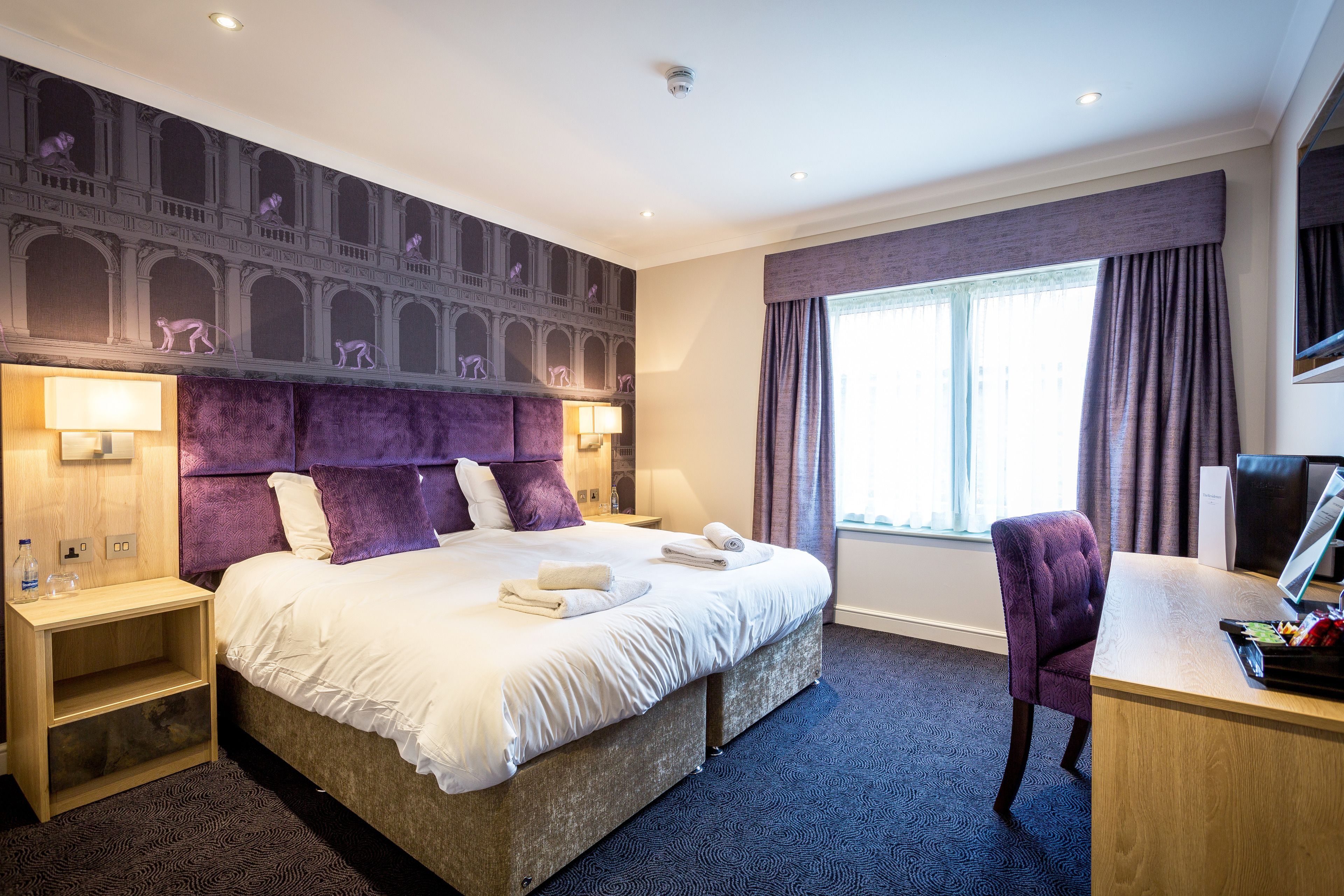 A hotel room with a large bed, a desk, a chair, a nightstand, and a wall mural.