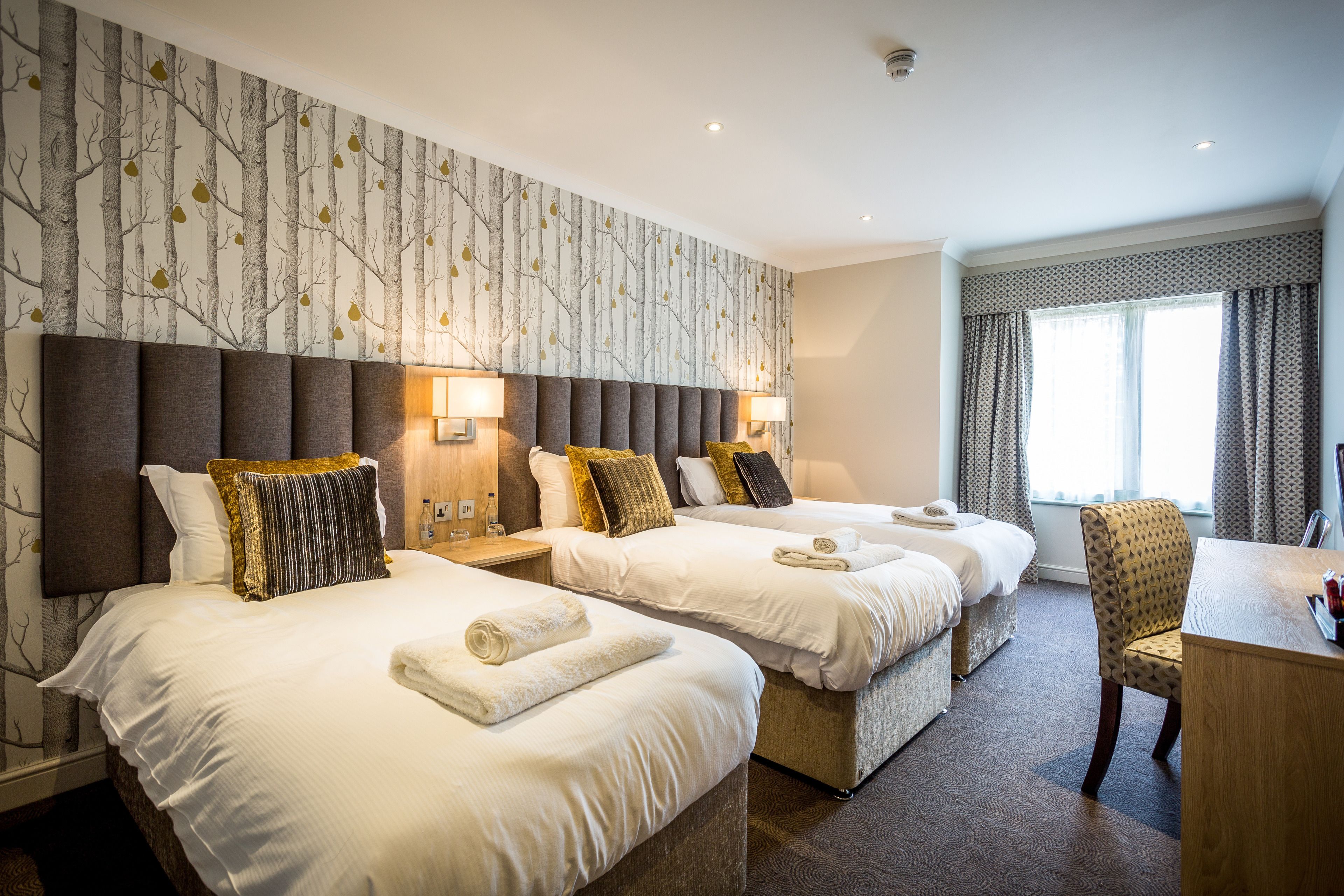 A hotel room with two beds, a desk, and a chair. There are lamps on the nightstands, and the room has a large window with curtains.