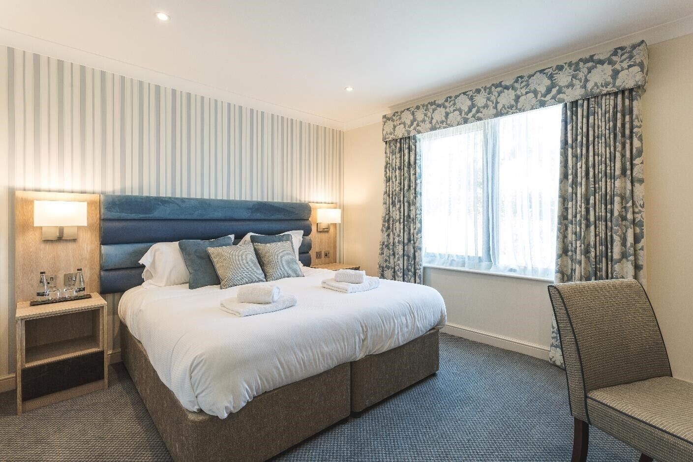 Een hotelkamer met een groot bed, twee nachtkastjes, een stoel en een raam met gordijnen.
