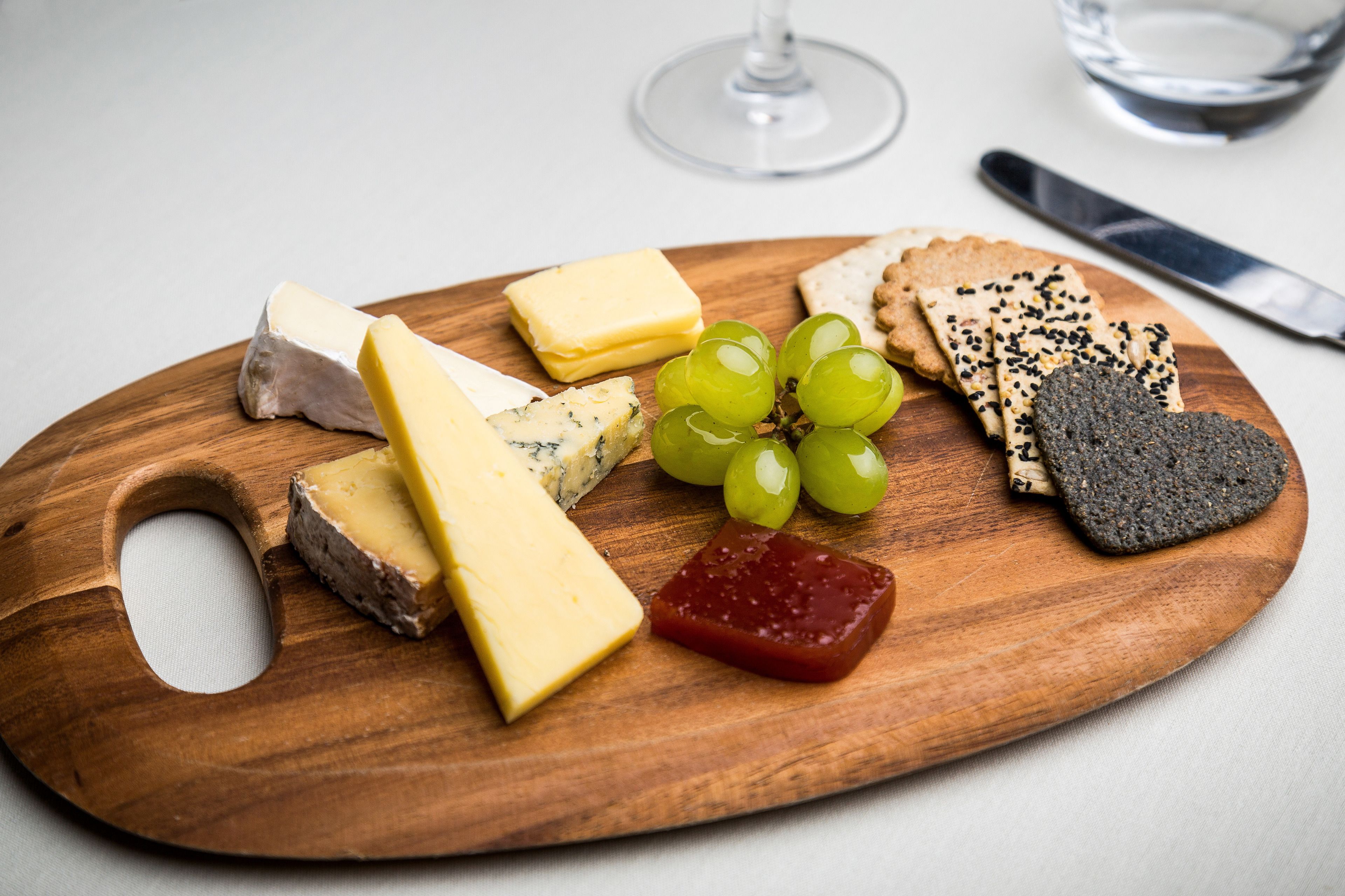 A wooden board with various cheeses, grapes, crackers, and a glass of wine.