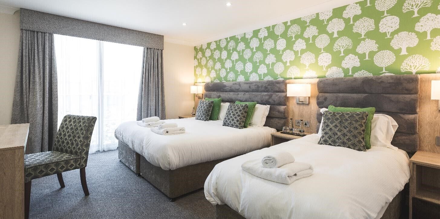 Quarto de hotel com duas camas, uma cadeira e uma janela grande com cortinas.