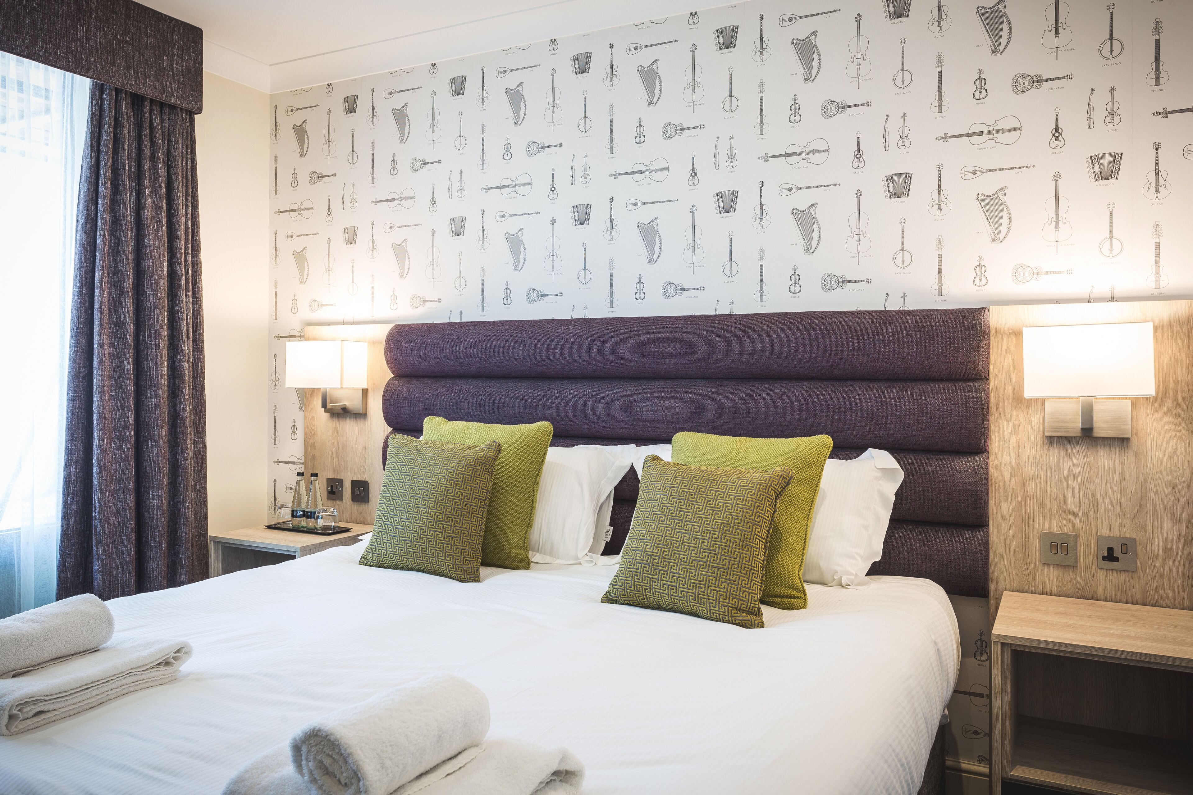 Habitación de hotel con una cama grande, dos almohadas, cabecera, lámparas de noche y una pared con motivos de instrumentos musicales.