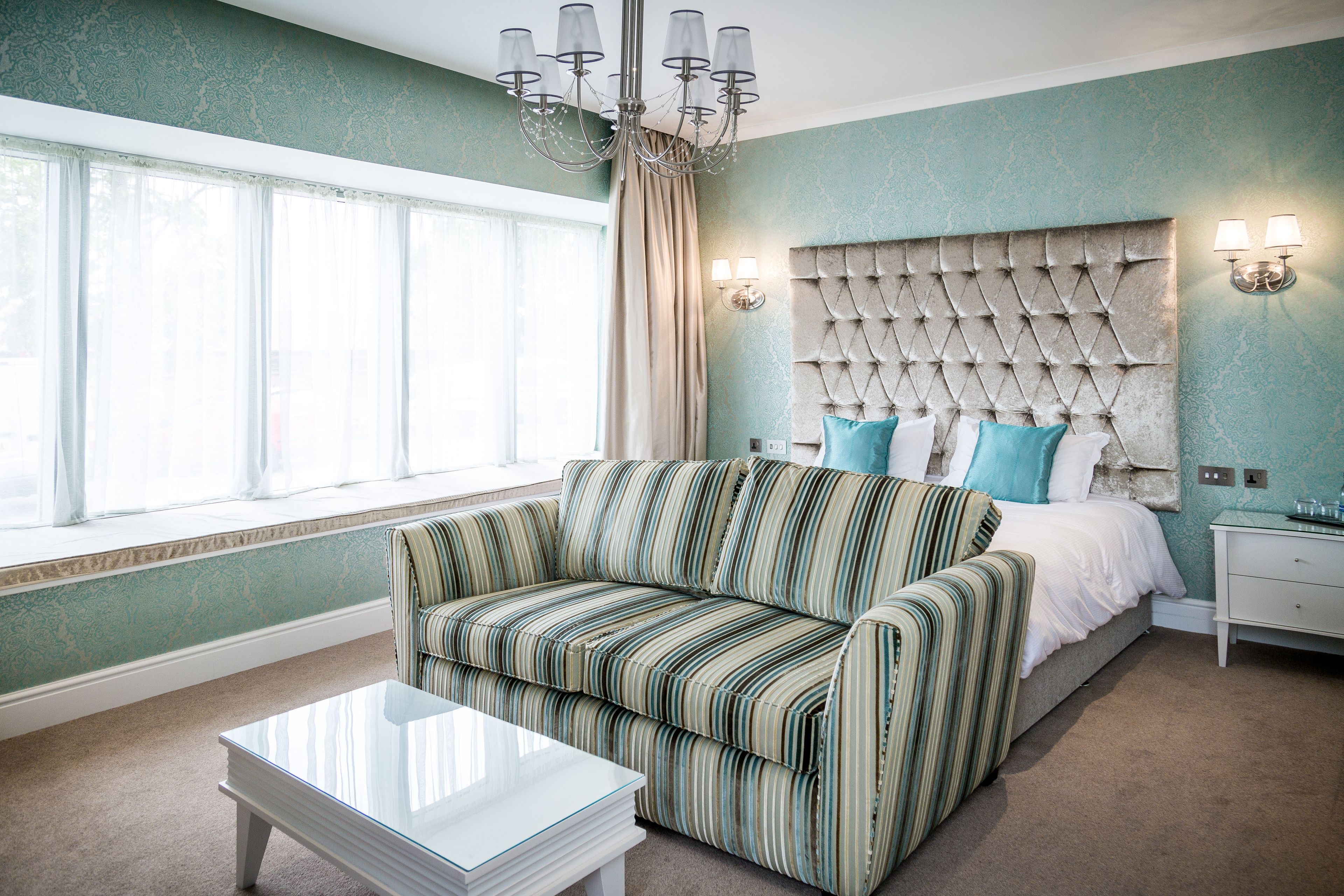 A bedroom with a tufted headboard, a striped sofa, a glass coffee table, and a window with curtains.