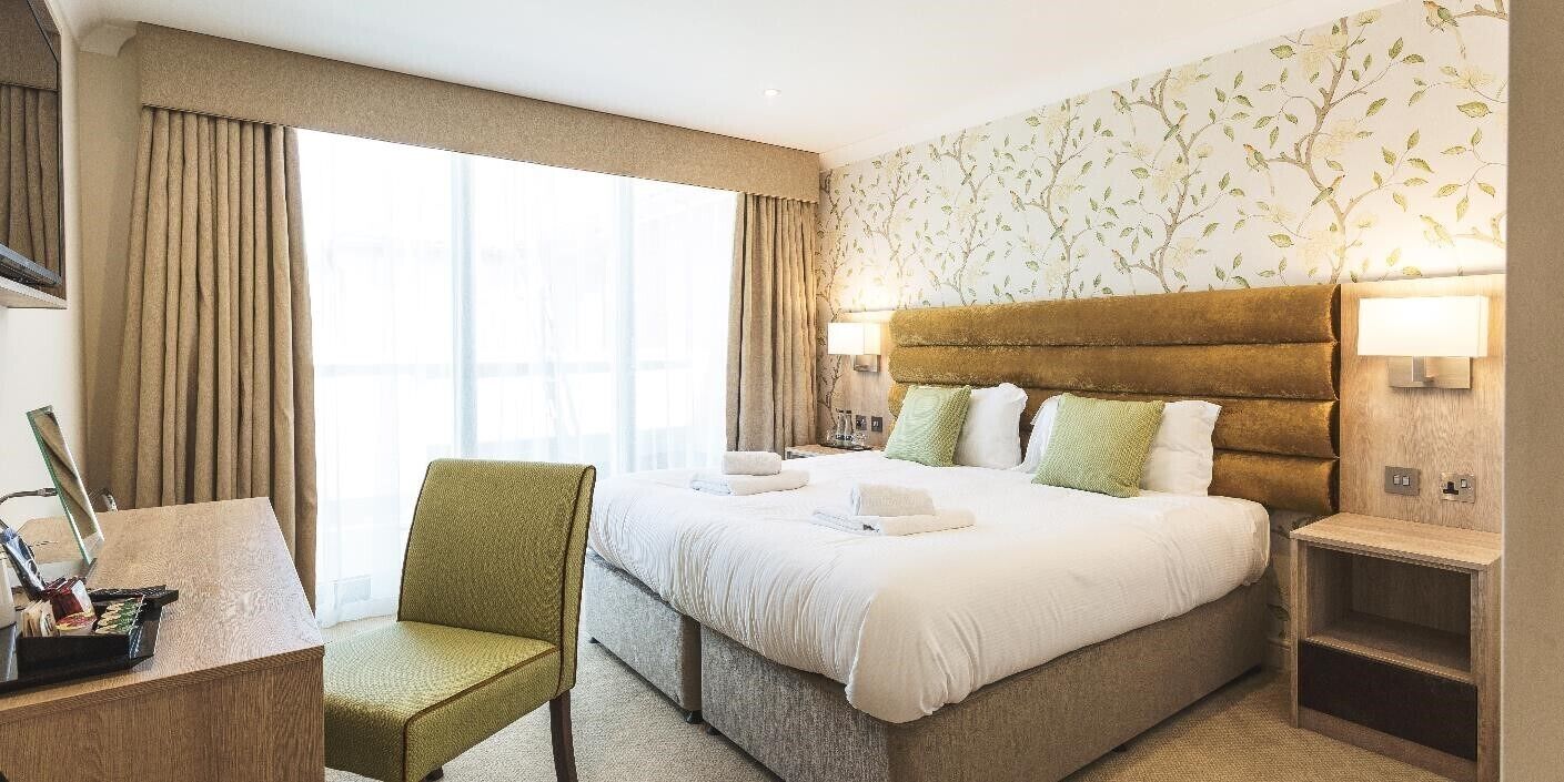 A hotel room with a large bed, a desk with a chair, and a view of the city through large windows.