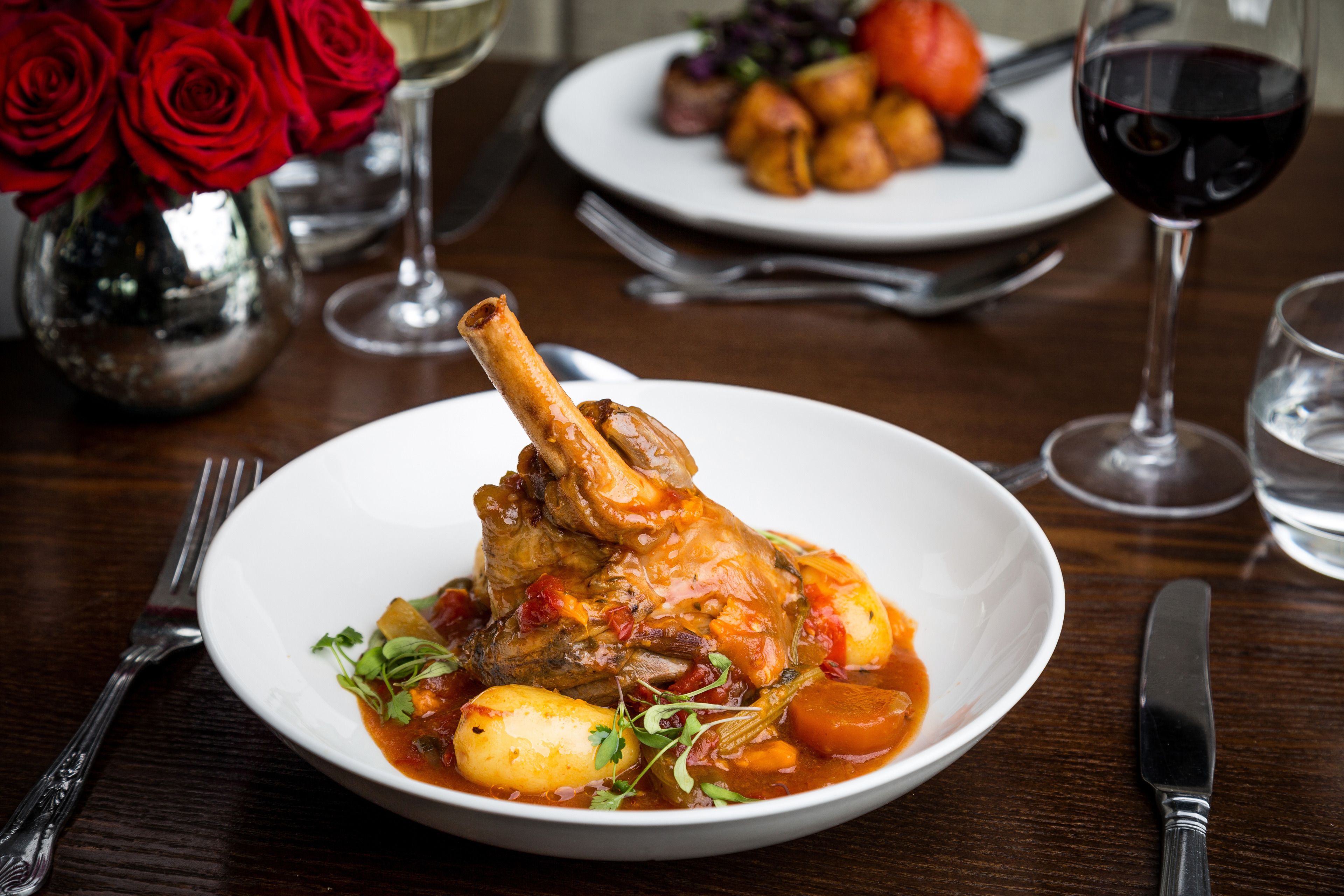 A dining table with a plate of food, a vase of red roses, and wine glasses.