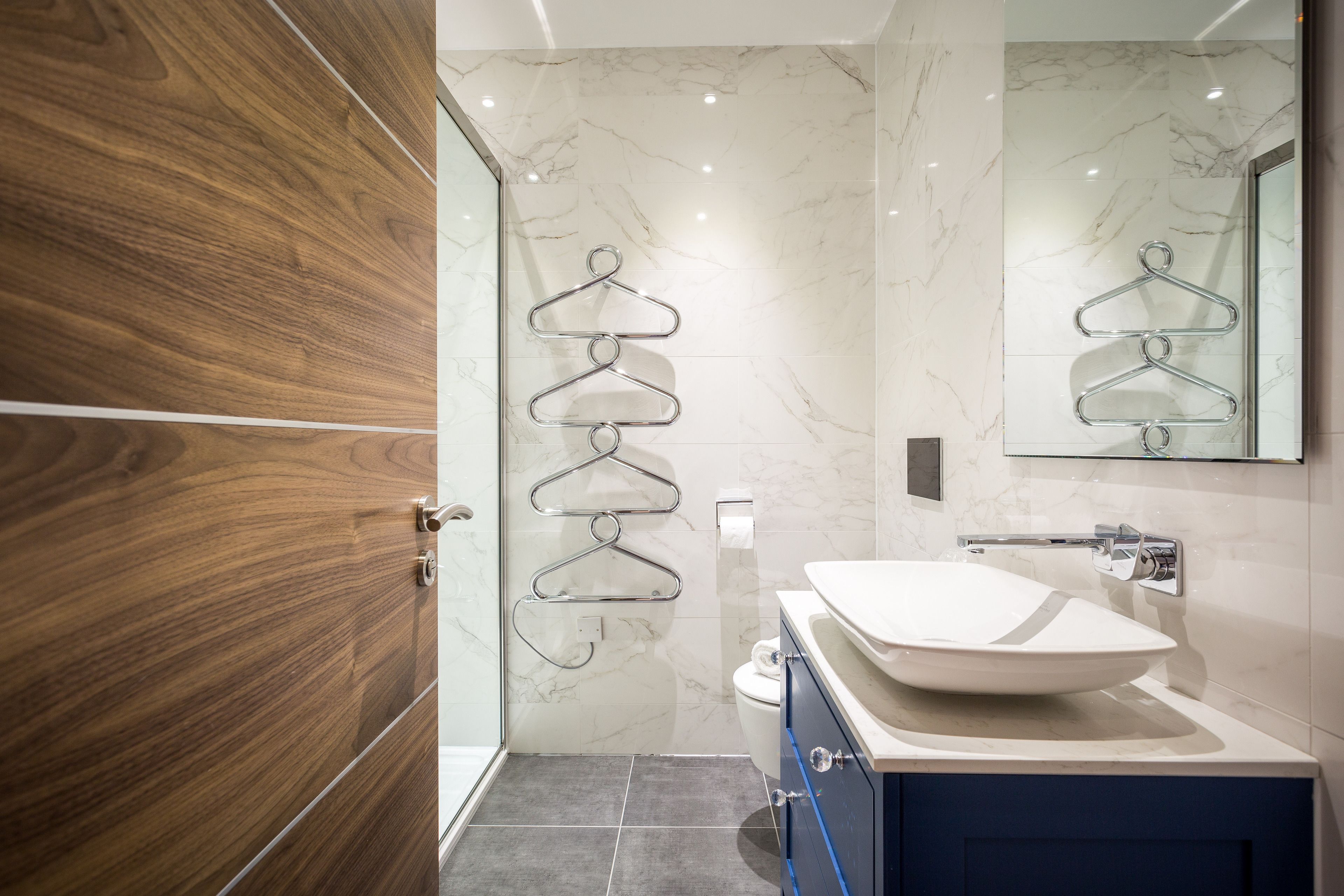 Un baño moderno con puerta de madera, pared de mármol, lavabo blanco y toallero.
