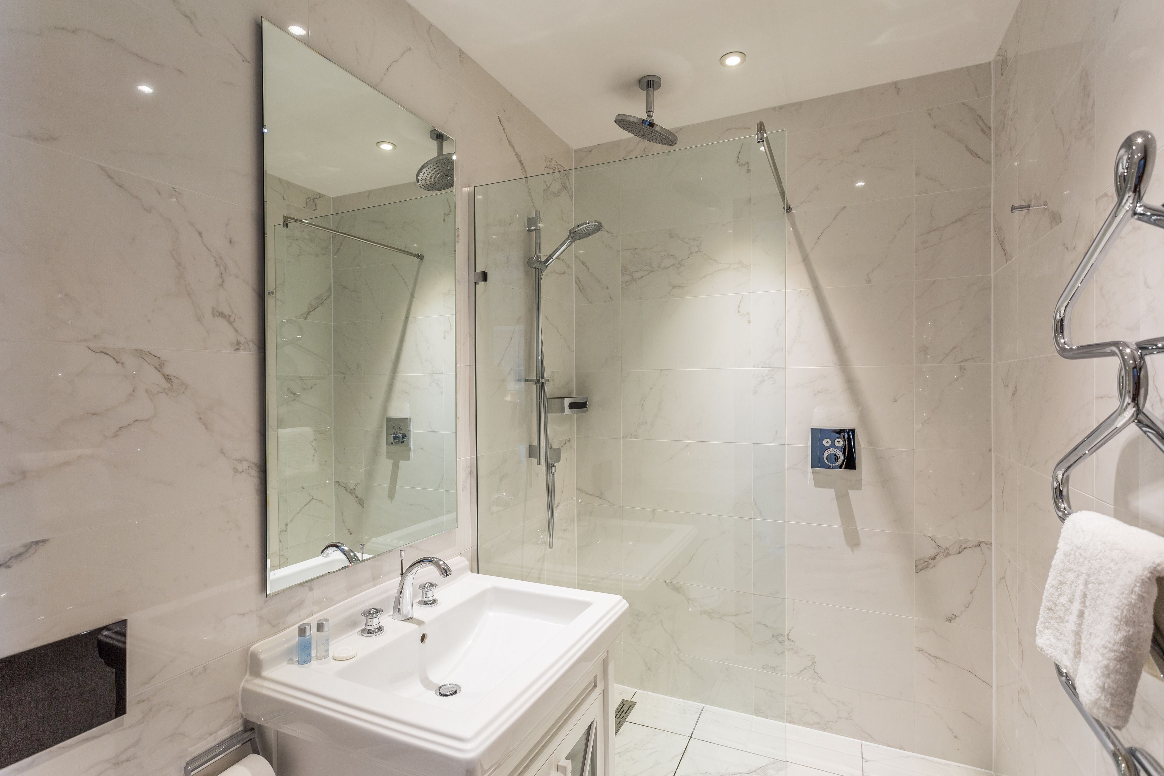 A modern bathroom with a marble wall, a large mirror, a sink, a glass-enclosed shower, and a towel rack.