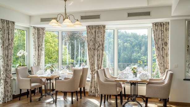 Comedor con mesa preparada para cuatro personas, ventanas amplias con cortinas y vista a zona boscosa.
