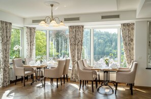A dining area with a table set for four, large windows with curtains, and a view of a forested area. - Romantikhotel Störmann (Schmallenberg)