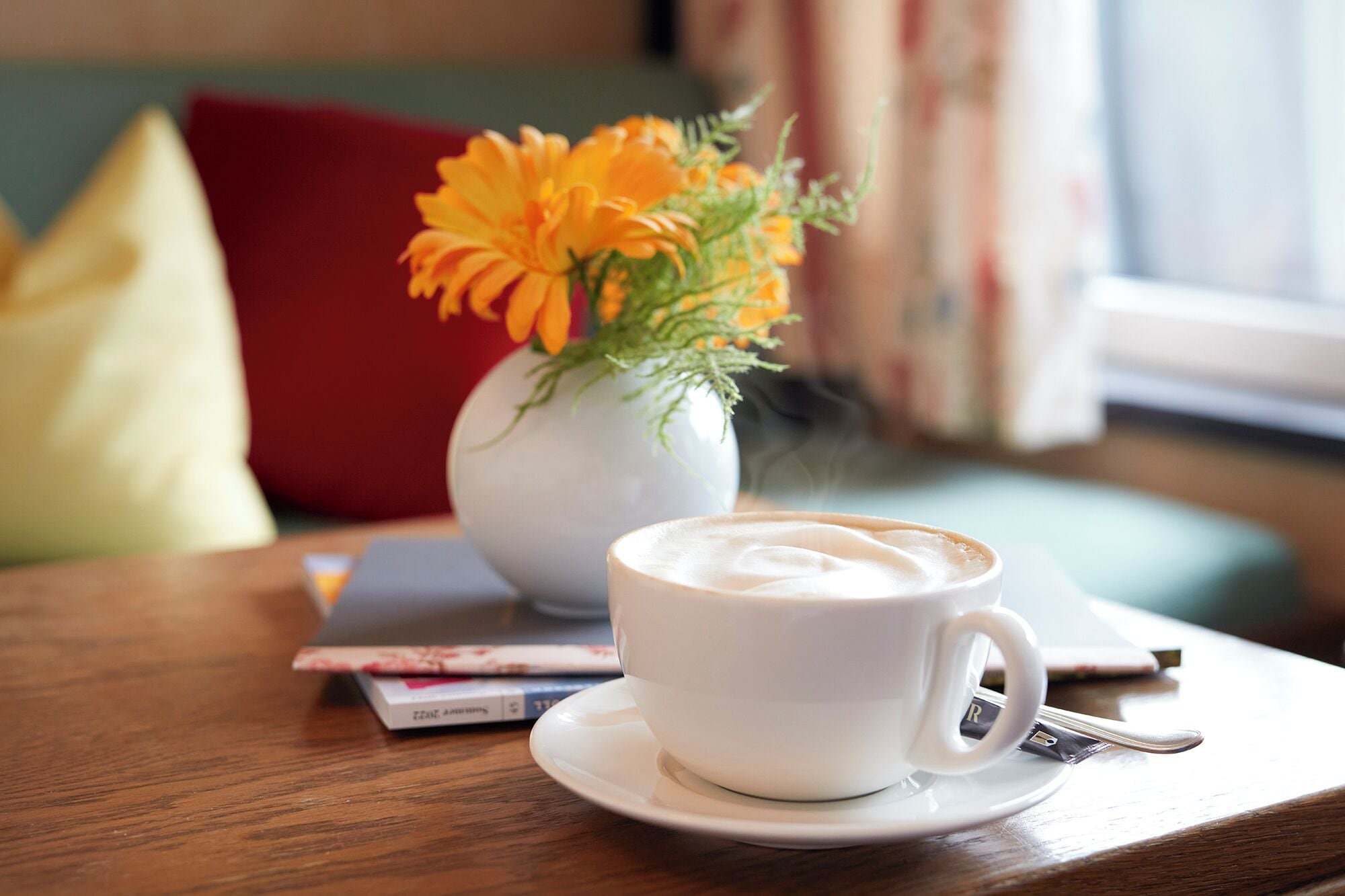 Eine weiße Kaffeetasse auf einem Untertasse mit einem Löffel, ein Buch und eine Vase mit orangefarbenen Blumen auf einem Holztisch.