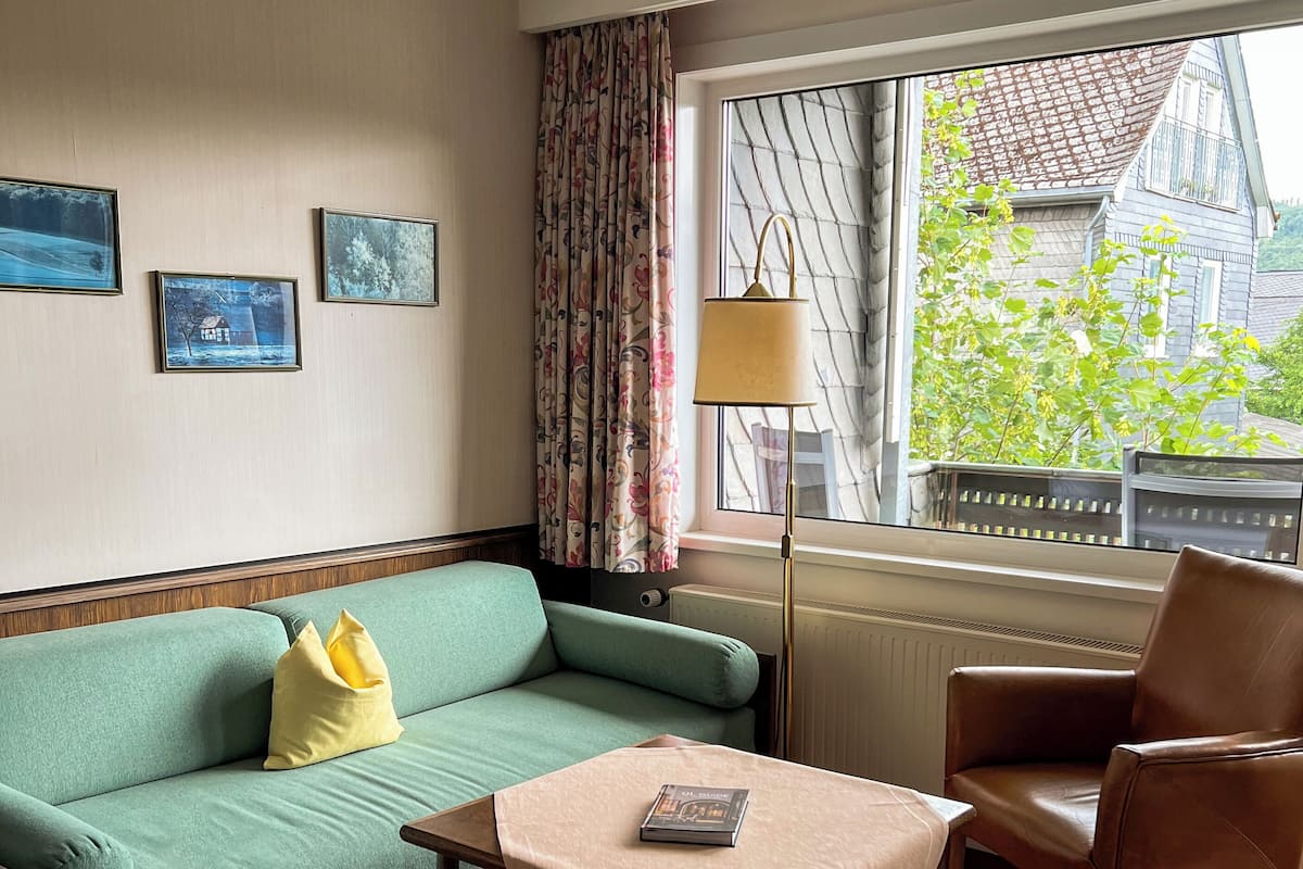 Habitación de hotel con sofá verde, una mesita de centro de madera, sillón de cuero marrón y ventana con vistas a edificios y áreas verdes.