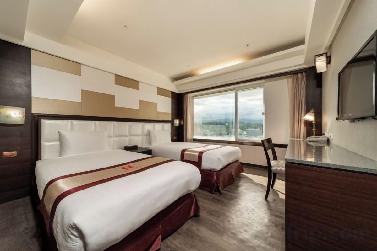 A hotel room with two beds, a desk, a chair, a TV, and a large window with a view.