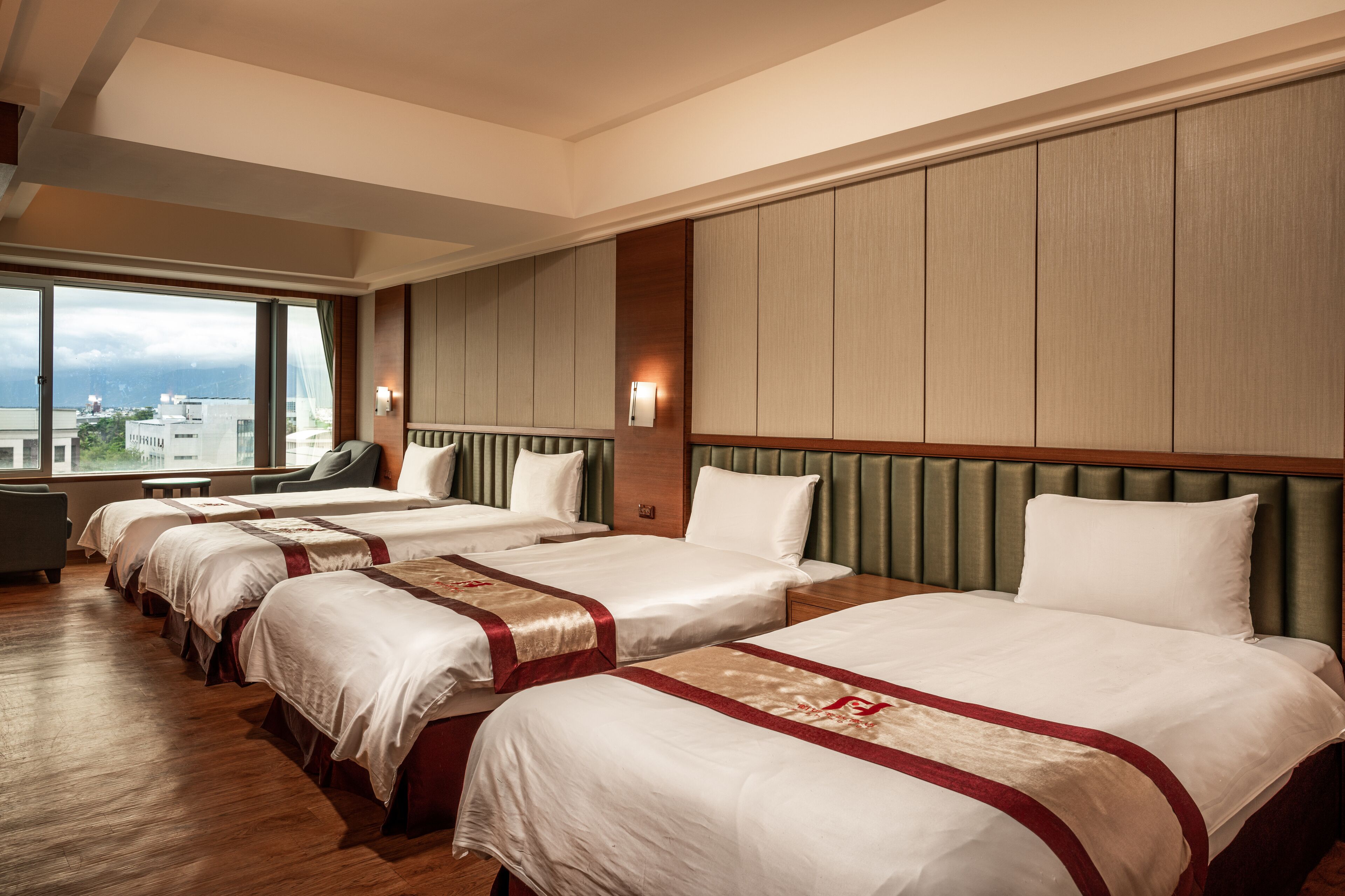 Une chambre d’hôtel avec quatre lits disposés en rangée, une grande fenêtre donnant sur la ville et une tête de lit recouverte d’un tissu vert.