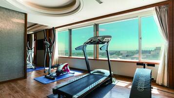 A modern hotel room with a treadmill, exercise equipment, and a large window offering a city view.