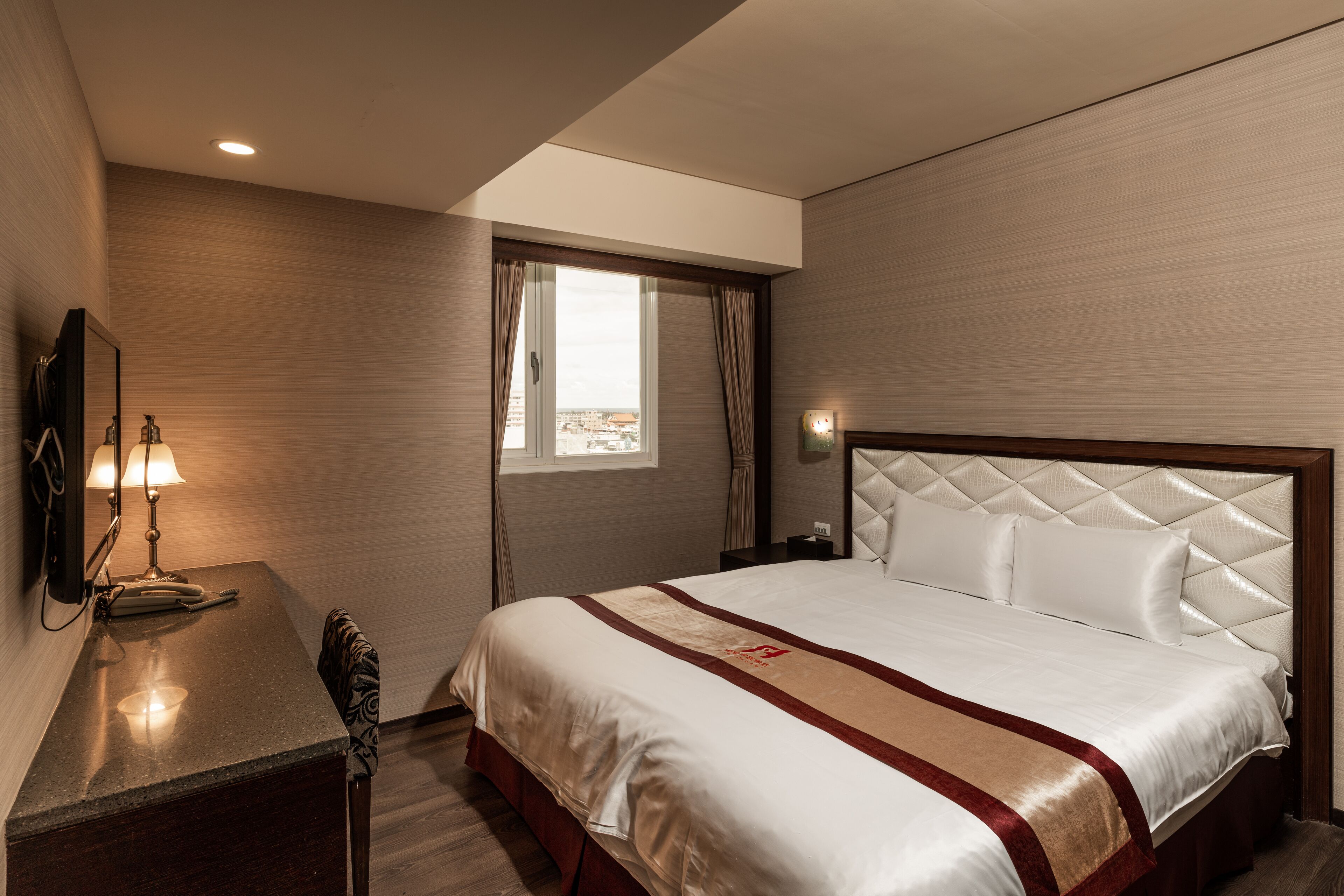 A hotel room with a large bed, a desk with a lamp, a window with curtains, and a view of the city.
