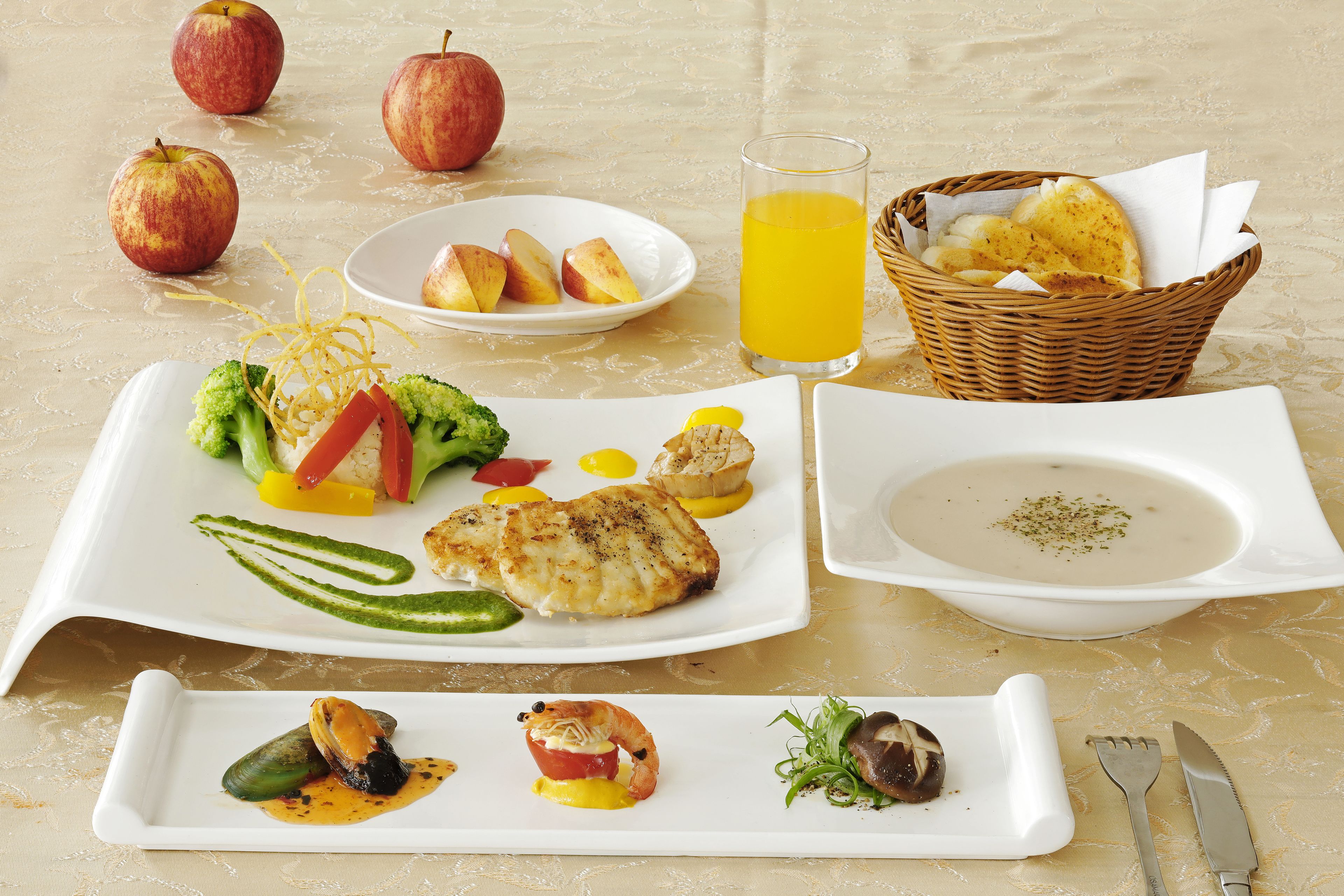 A table set with a variety of dishes including seafood, vegetables, and fruits, along with a glass of orange juice and a basket of bread.
