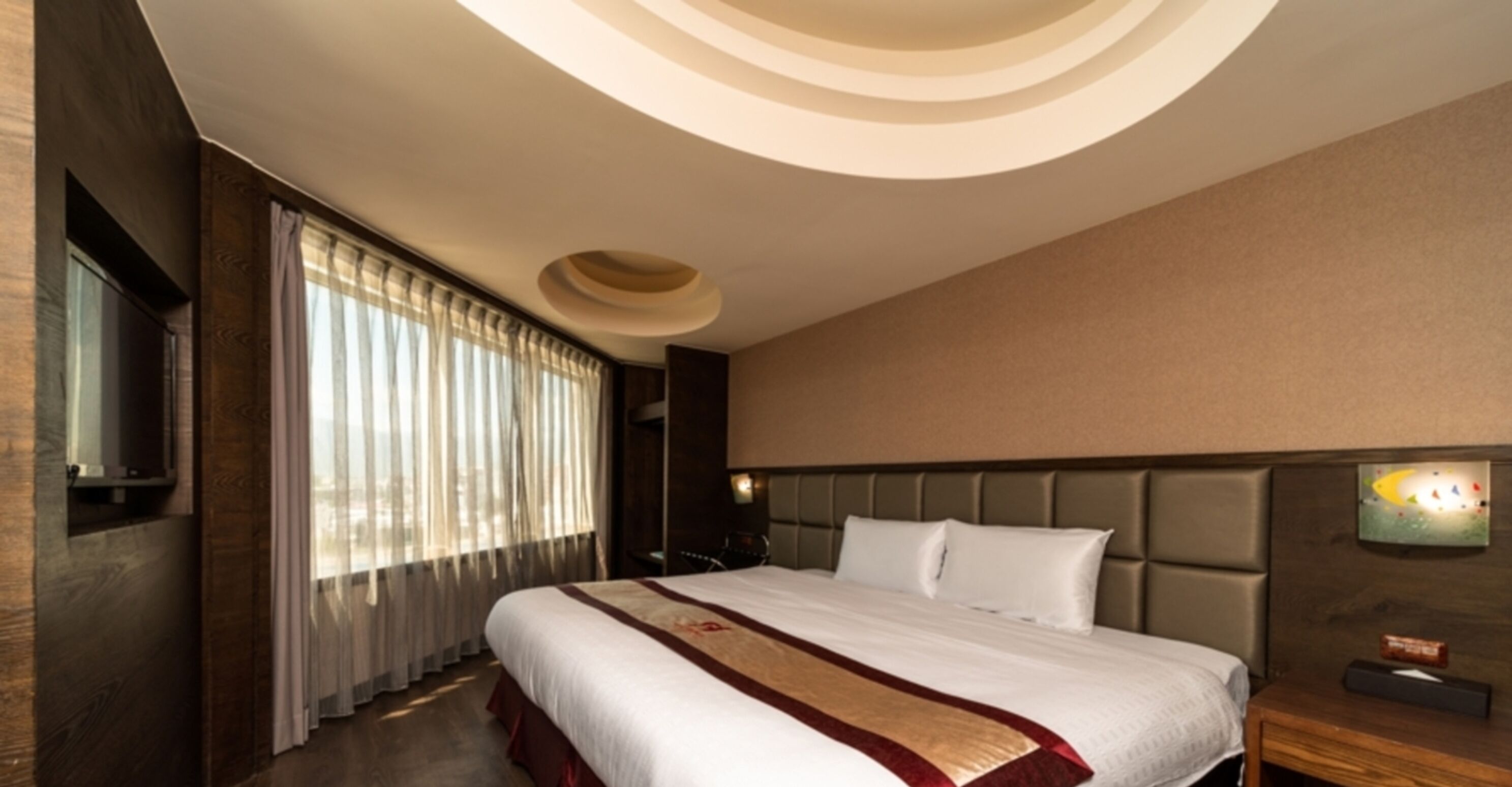 A hotel room with a large bed, a headboard with a light fixture, a bedside table, and a window with curtains.