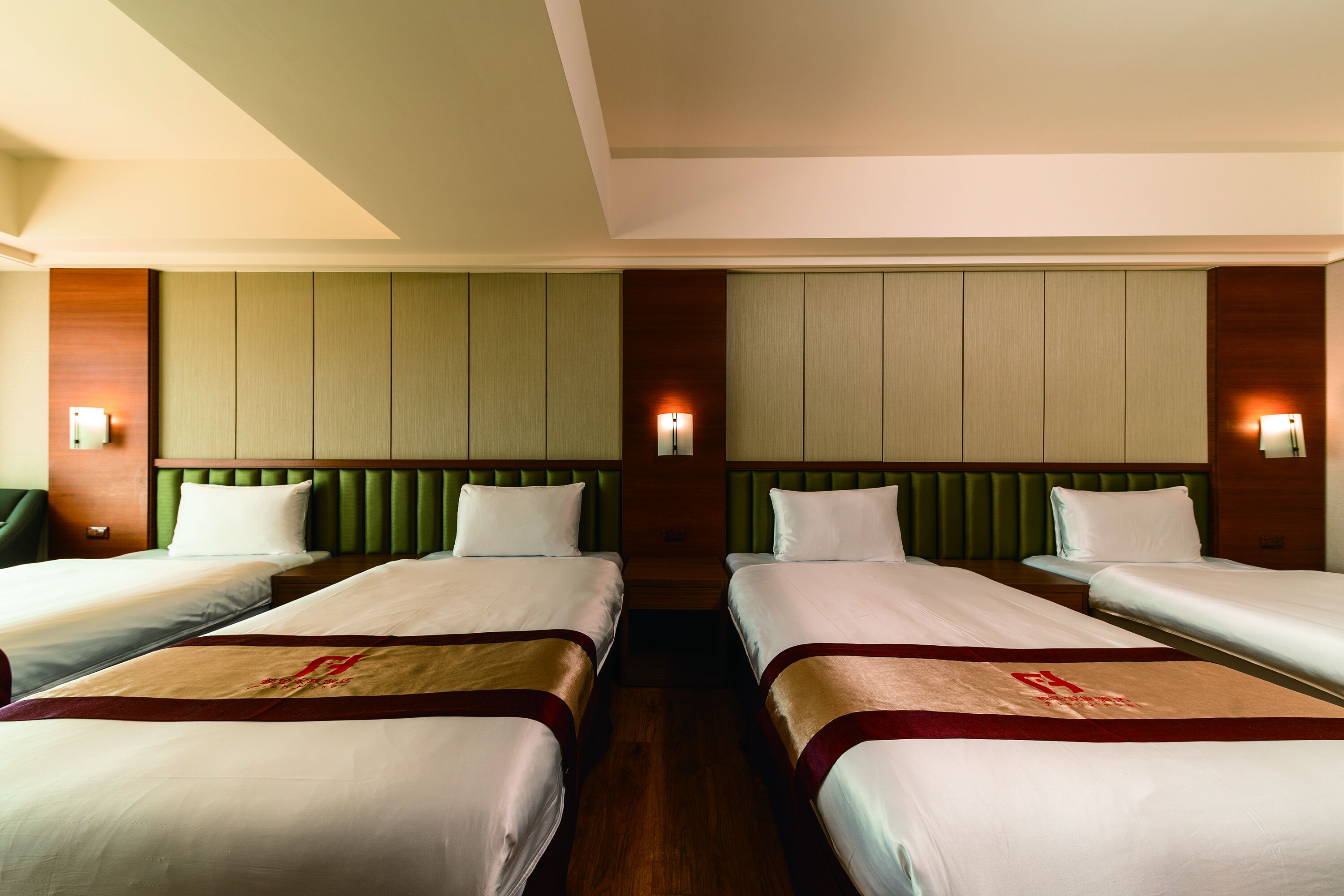 A hotel room with four single beds, green padded headboards, and wall-mounted lights.
