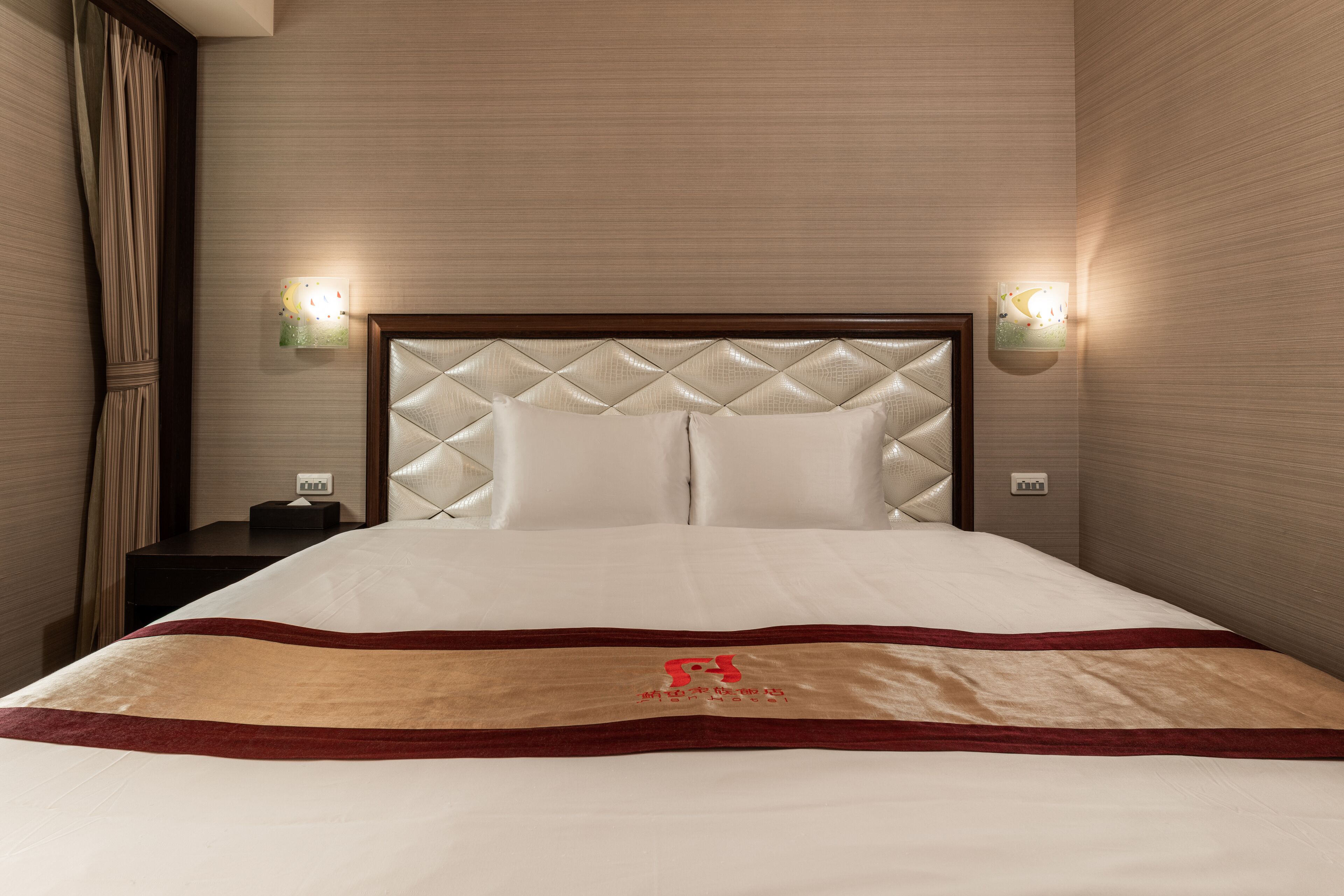 A hotel room with a large bed, a headboard with a diamond pattern, two wall-mounted light fixtures, and a bedside table with a lamp.