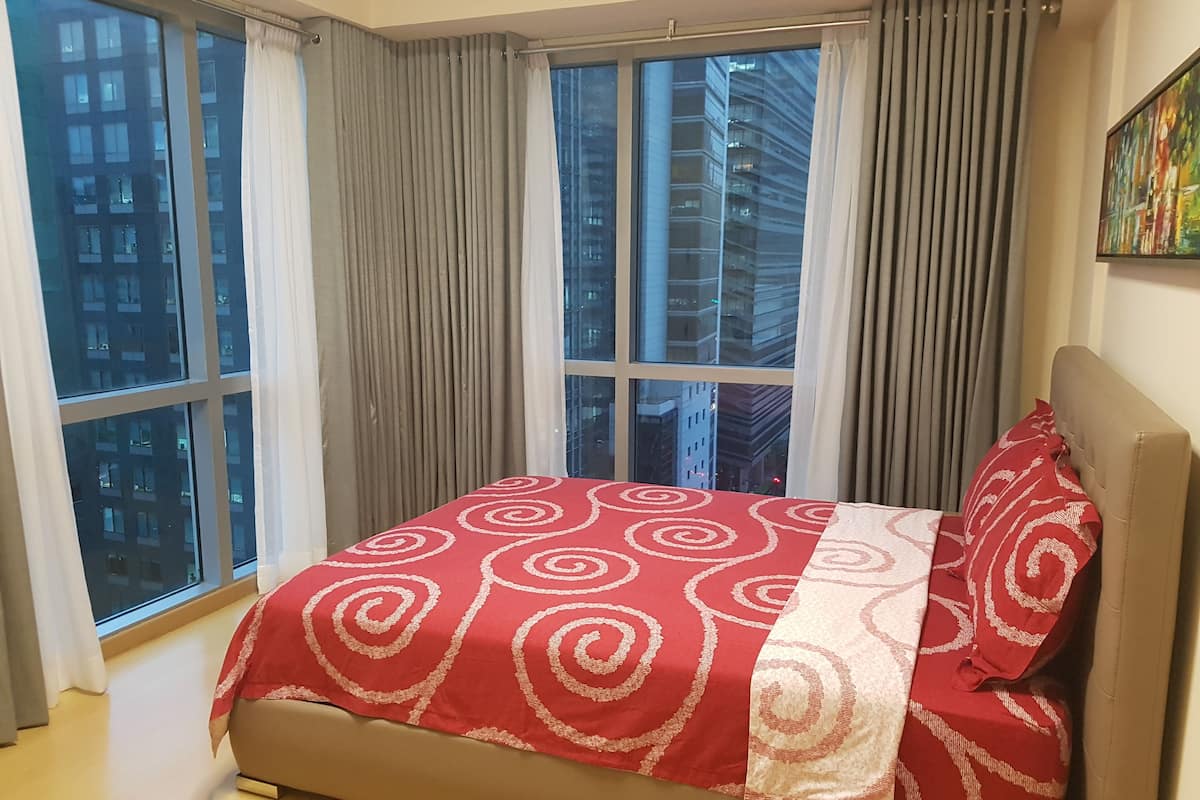 A bedroom with a large window, a bed with a red and white patterned bedspread, and a city view.