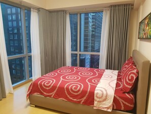 A bedroom with a large window, a bed with a red and white patterned bedspread, and a city view. - Avant Apartments At The Fort (Taguig)
