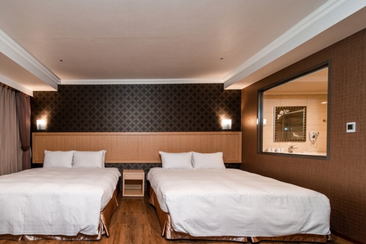 Habitación de hotel con dos camas, cabecera de madera, un espejo grande y un baño visible a través de una puerta abierta.