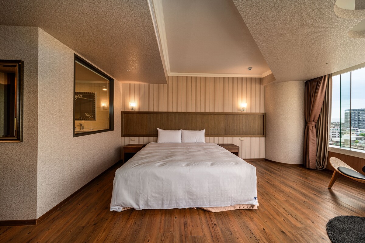 Una habitación de hotel moderna con una cama grande, suelo de madera, un espejo amplio y vistas a la ciudad.