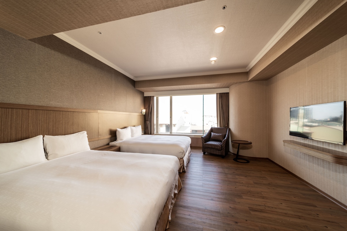 Habitación de hotel con dos camas, televisión, una silla y ventana con cortinas.