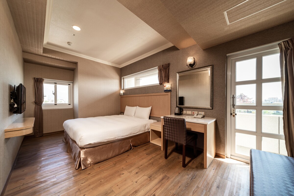 Habitación de hotel con cama, escritorio, silla y un ventanal grande.