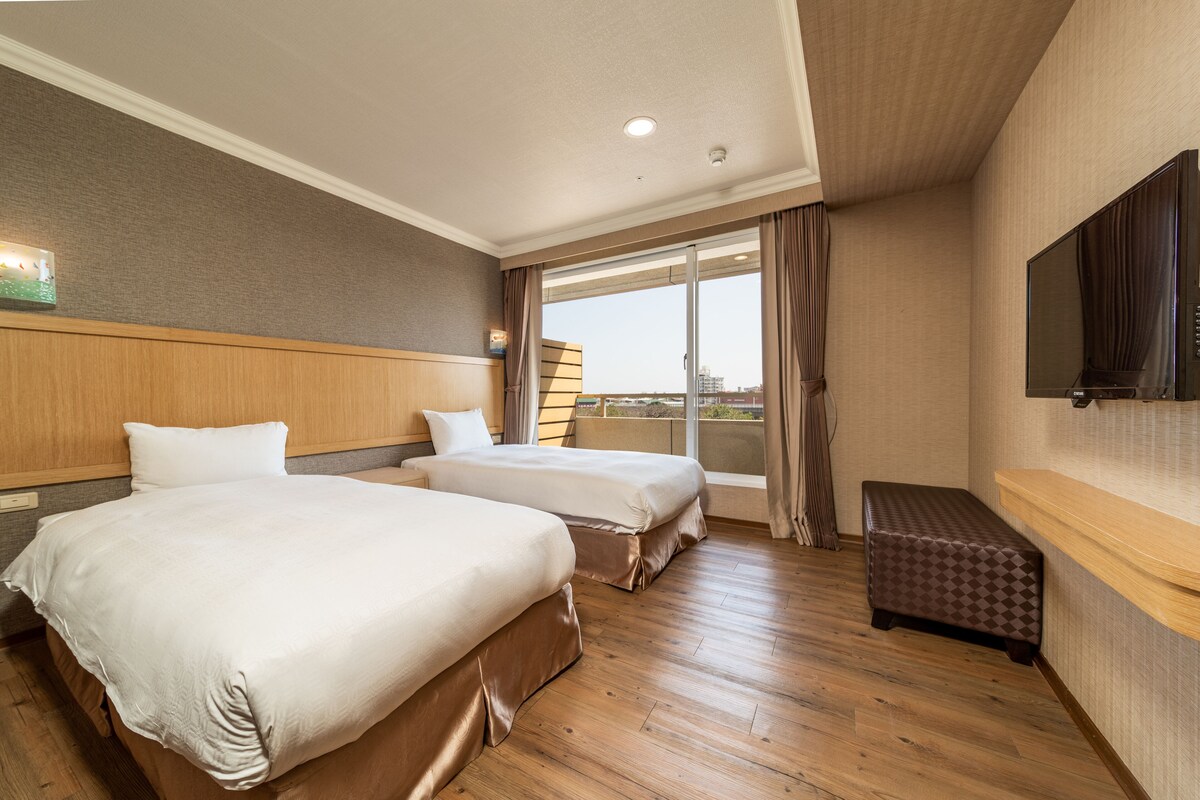 Habitación de hotel con dos camas, un banco de madera, televisión y un amplio ventanal con cortinas.