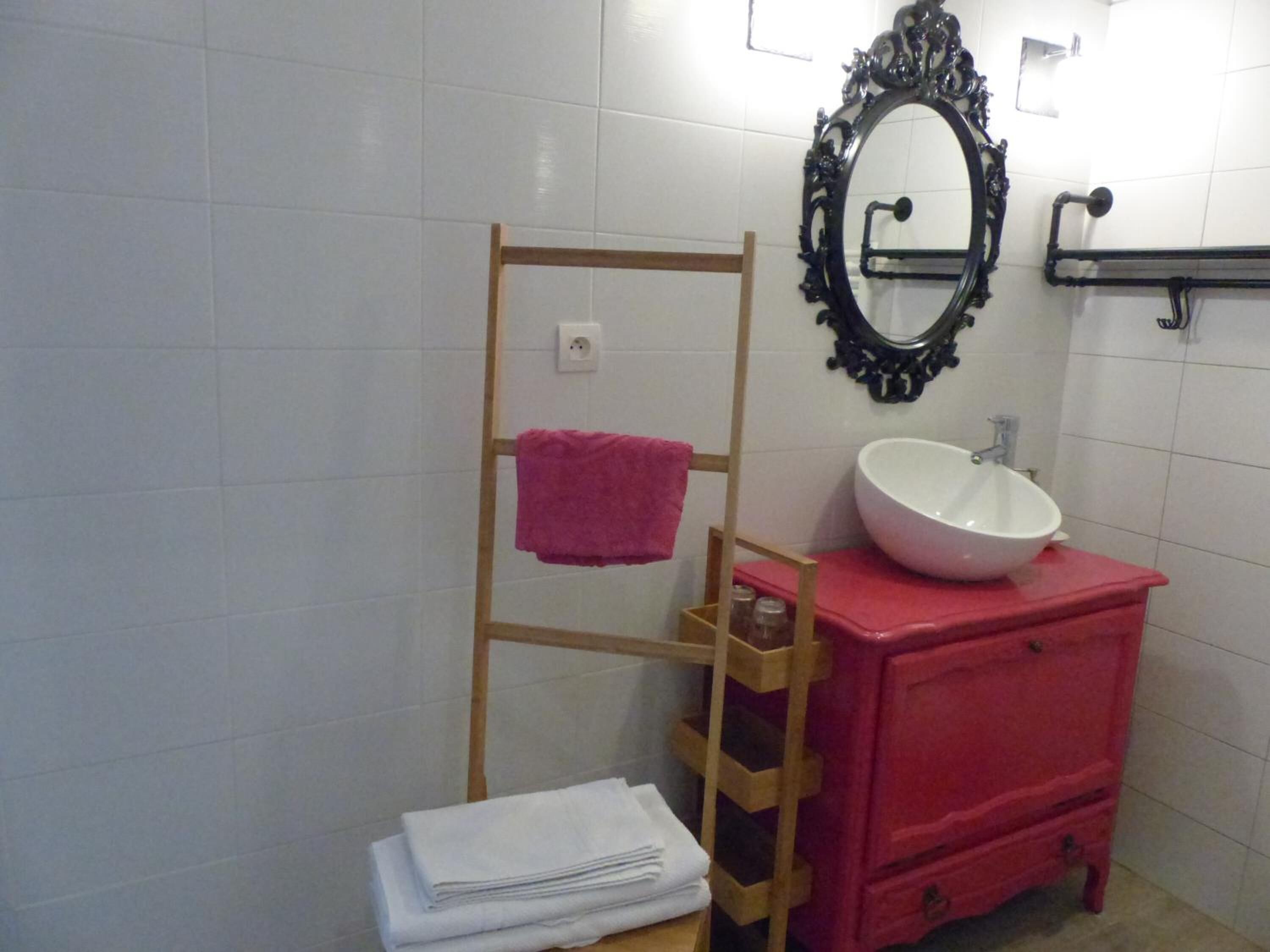 Ett badrum med vita, kaklade väggar, ett trähandduksställ med en rosa handduk, en vit tvättställ placerad på en röd bänkskiva och en svart, utsmyckad spegel.
