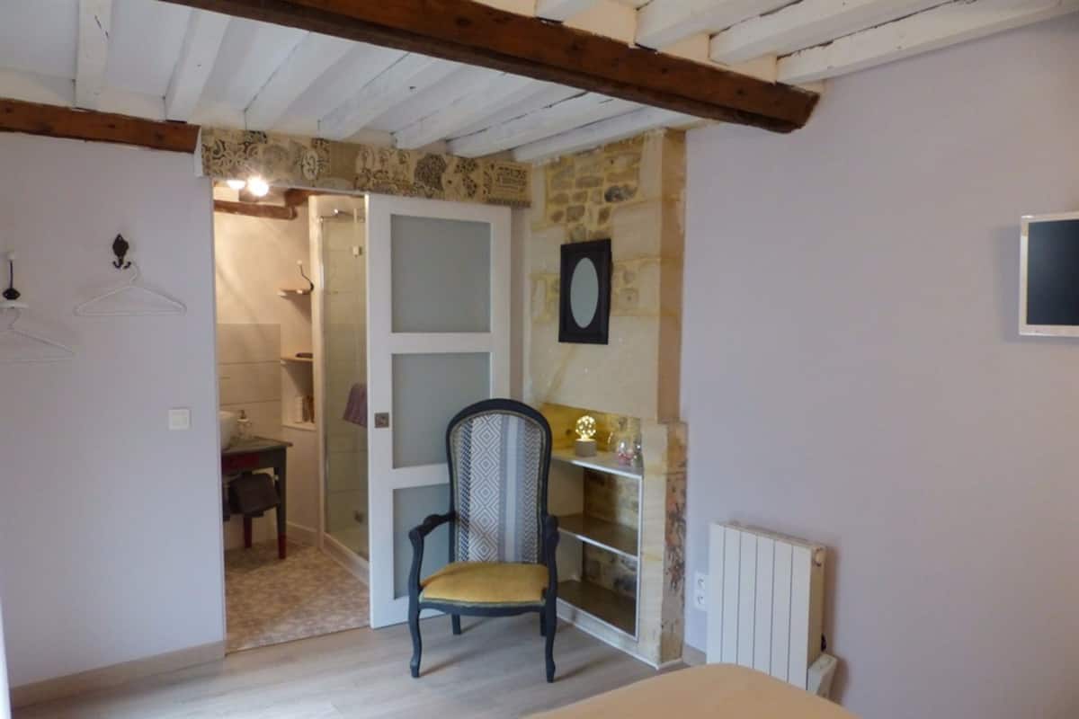 Quarto com cama, poltrona, prateleira com objetos decorativos, espelho e porta de acesso a um banheiro.