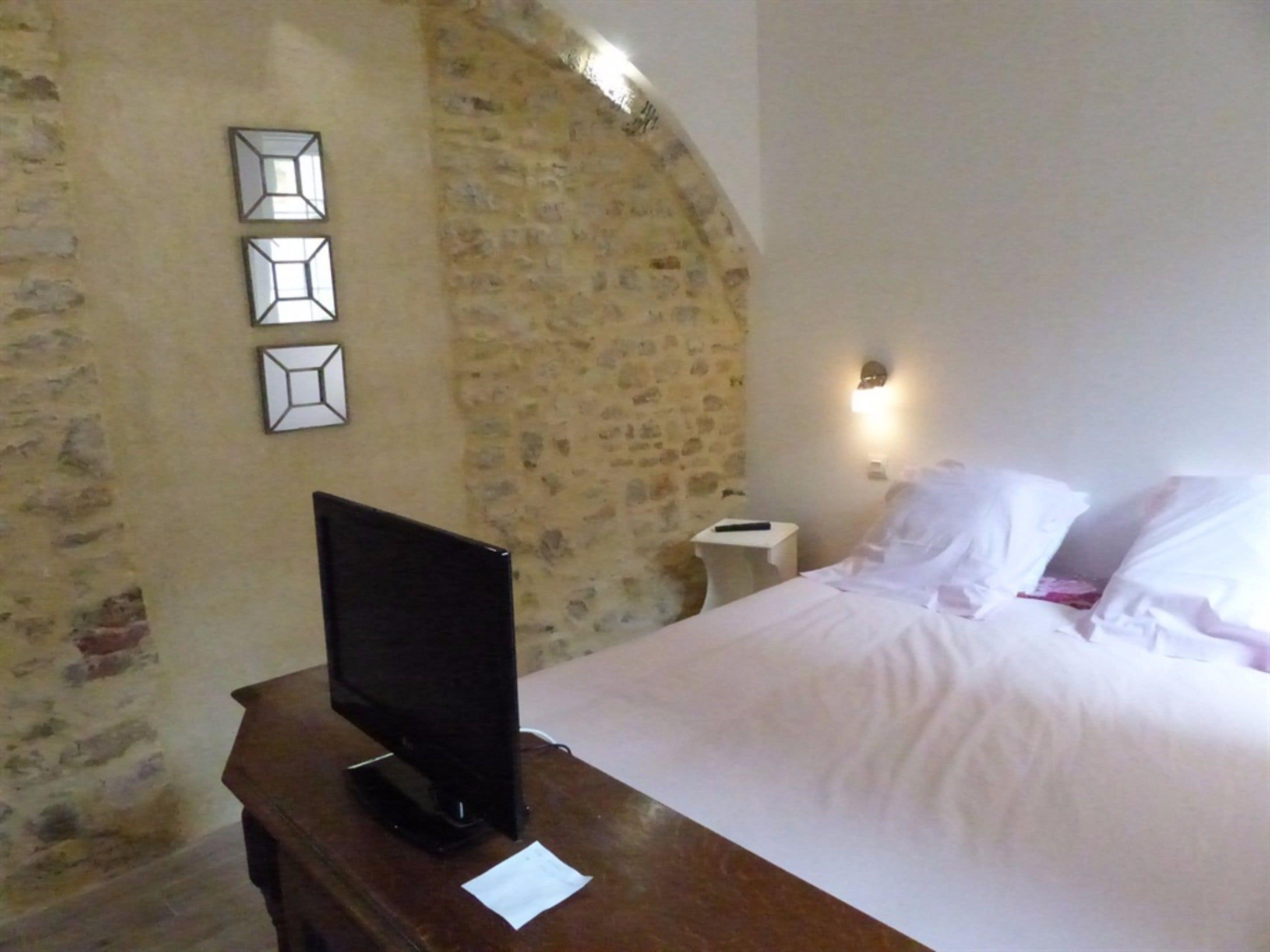 Een slaapkamer met een stenen muur, een bed met witte lakens, een nachtkastje met een afstandsbediening en een flatscreen televisie op een houten dressoir.