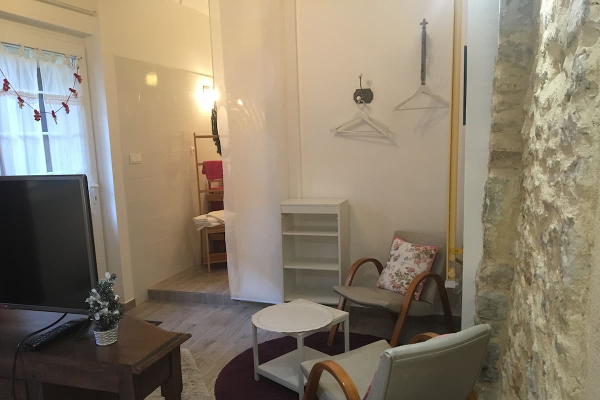 Um espaço compacto para viver, com televisão, uma mesinha, uma cadeira e um armário com prateleiras.