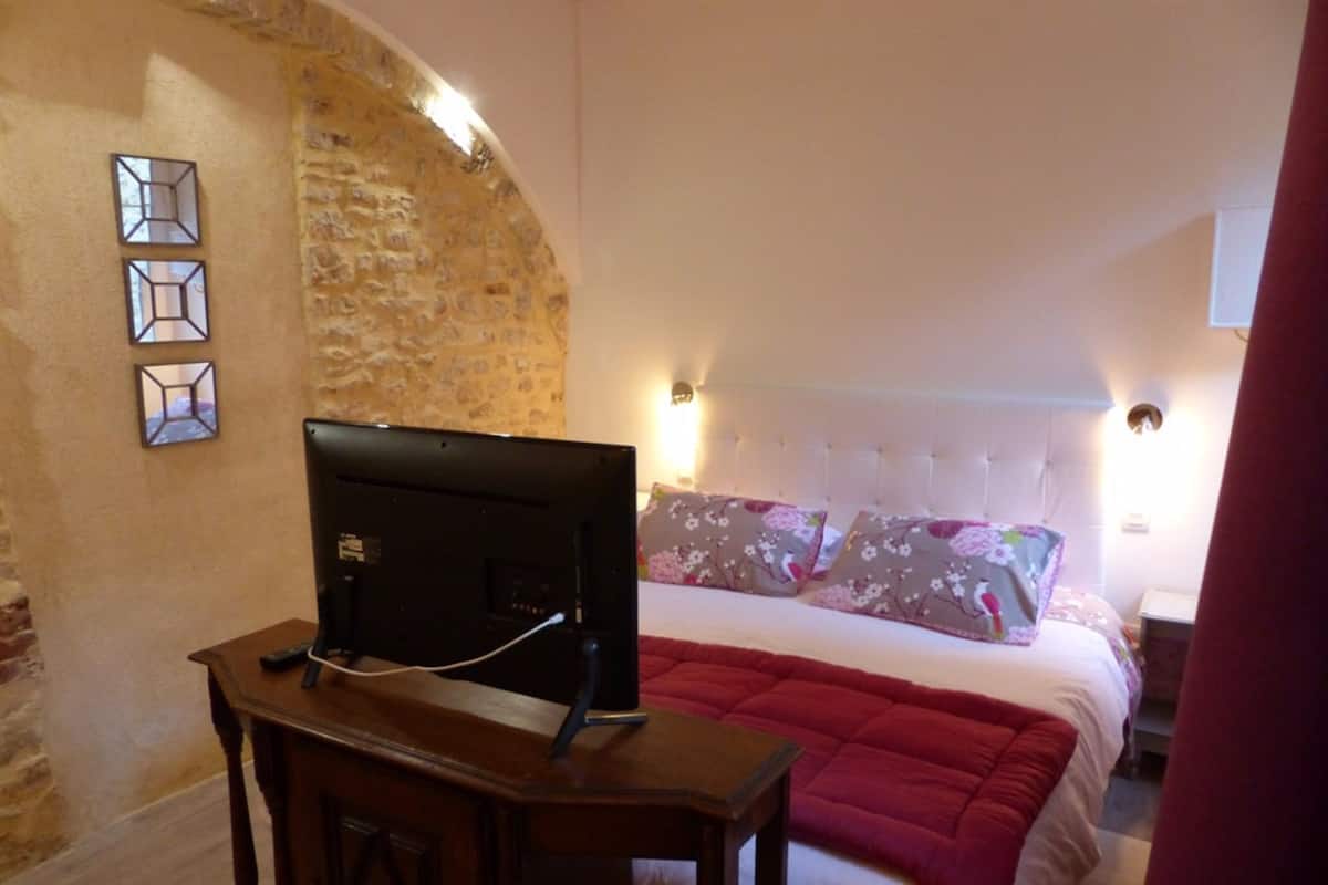 Quarto com parede de pedra, cama com almofadas florais e televisão sobre mesa de madeira.