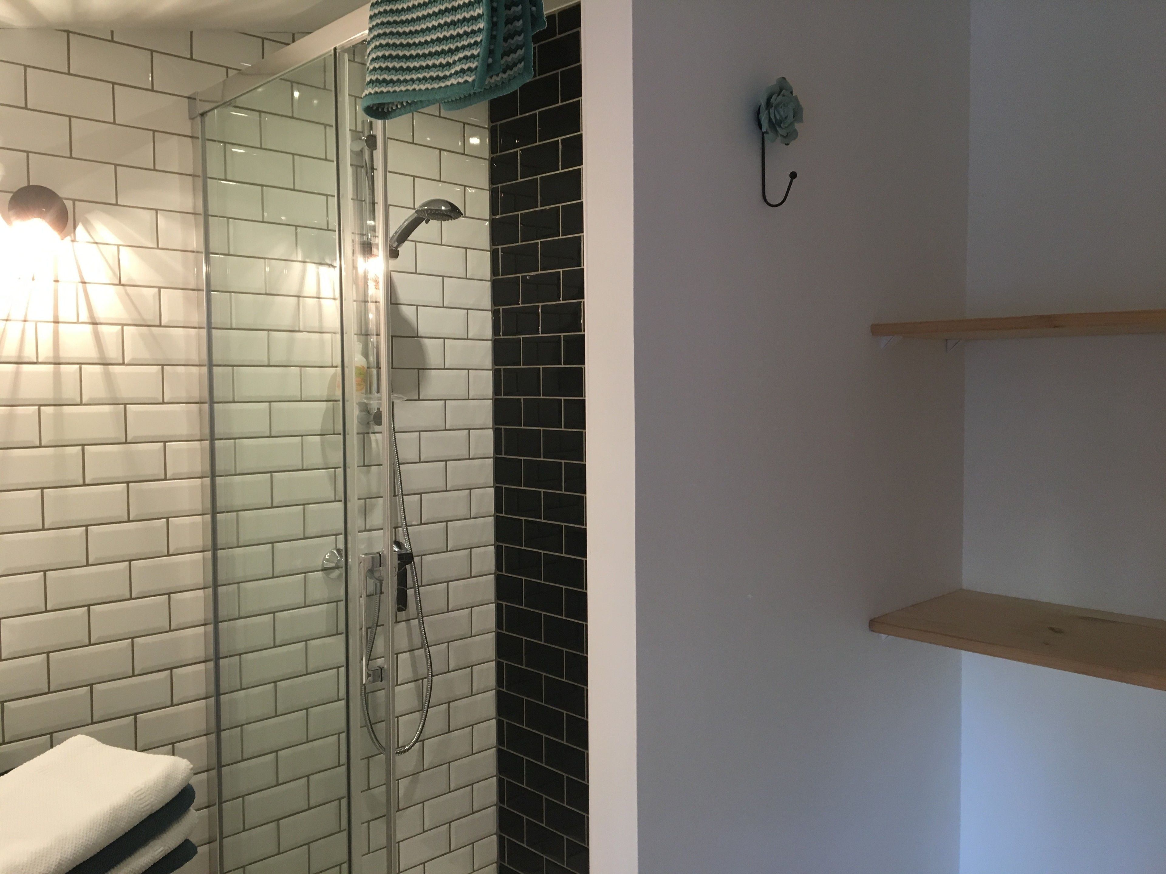 A bathroom with a glass shower enclosure, white and black tiled walls, a towel rack, and a wooden shelf.