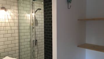 A bathroom with a glass shower enclosure, white and black tiled walls, a towel rack, and a wooden shelf.
