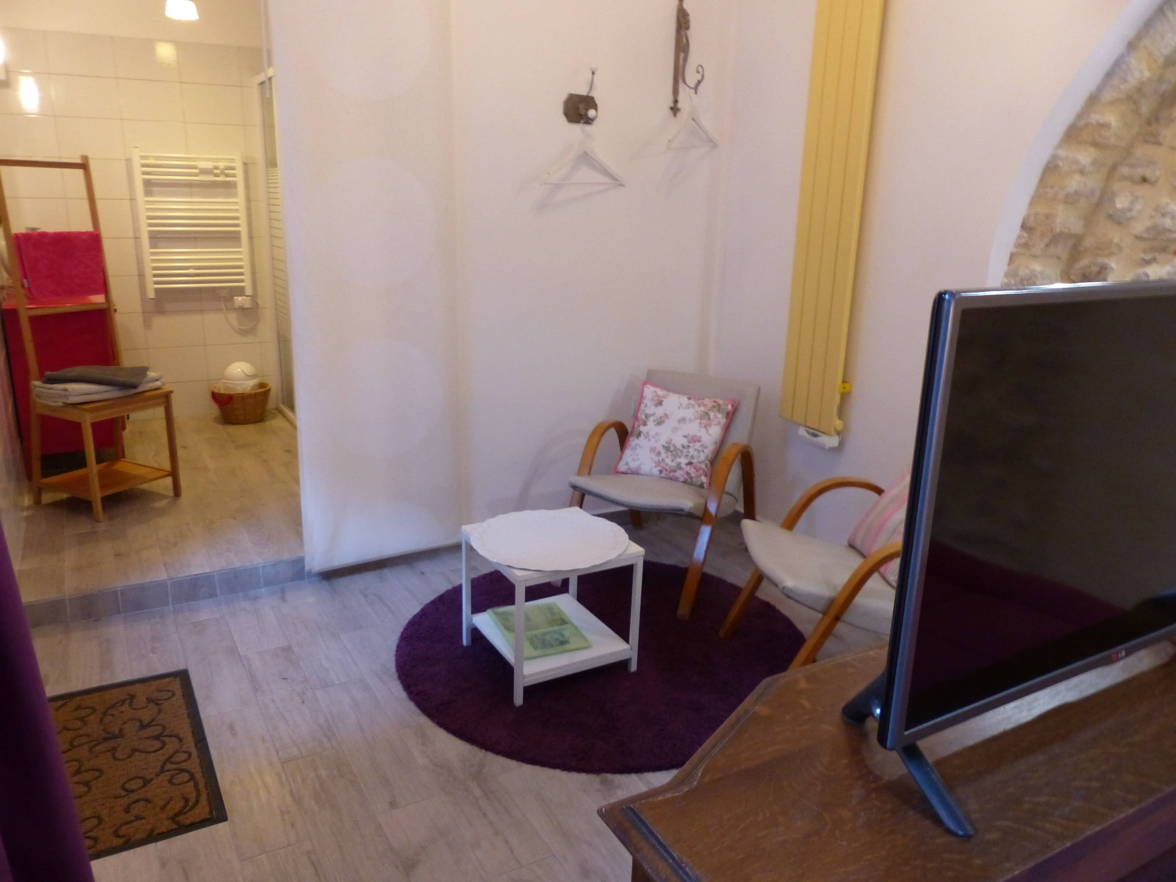 A small, cozy room with a flat-screen TV, a white table, and two chairs. There is a red towel rack and a basket on the floor.