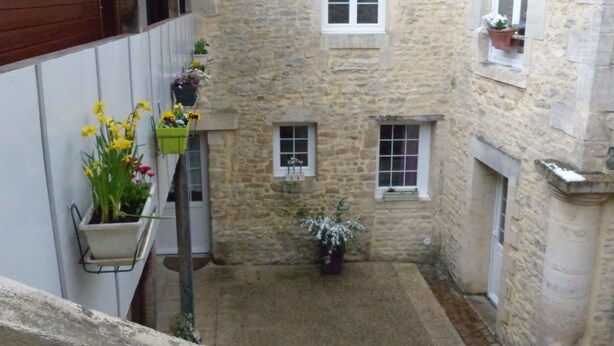 Une étroite cour pavée, avec une porte en bois sur la gauche, une petite fenêtre agrémentée d’une jardinière sur la droite et un pot de fleurs placé au centre.