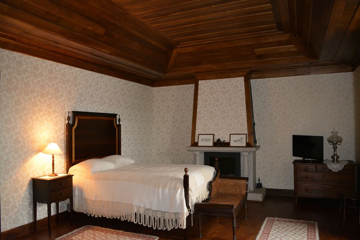 A bedroom with a wooden ceiling, a bed with a white bedspread, a nightstand with a lamp, a fireplace, and a television on a dresser.