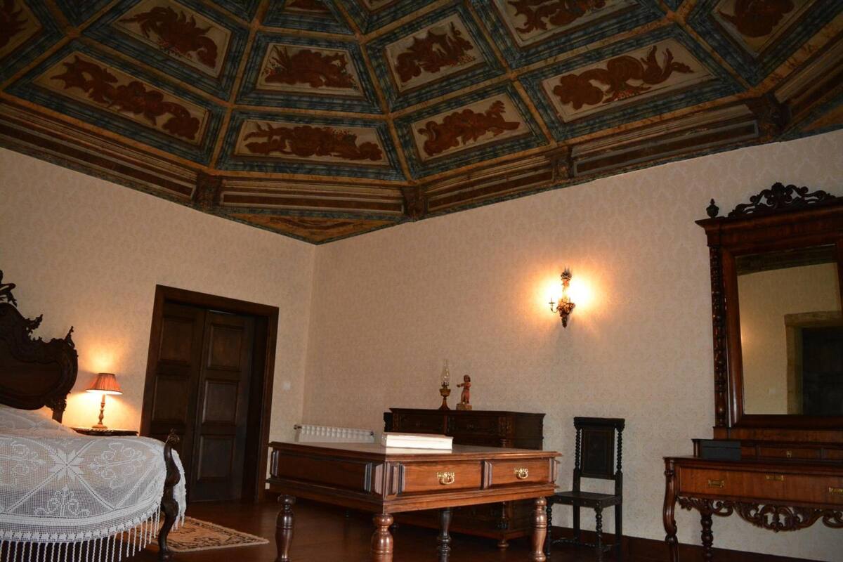 A room with a wooden bed, a desk, a chair, a large mirror, and a decorative ceiling.