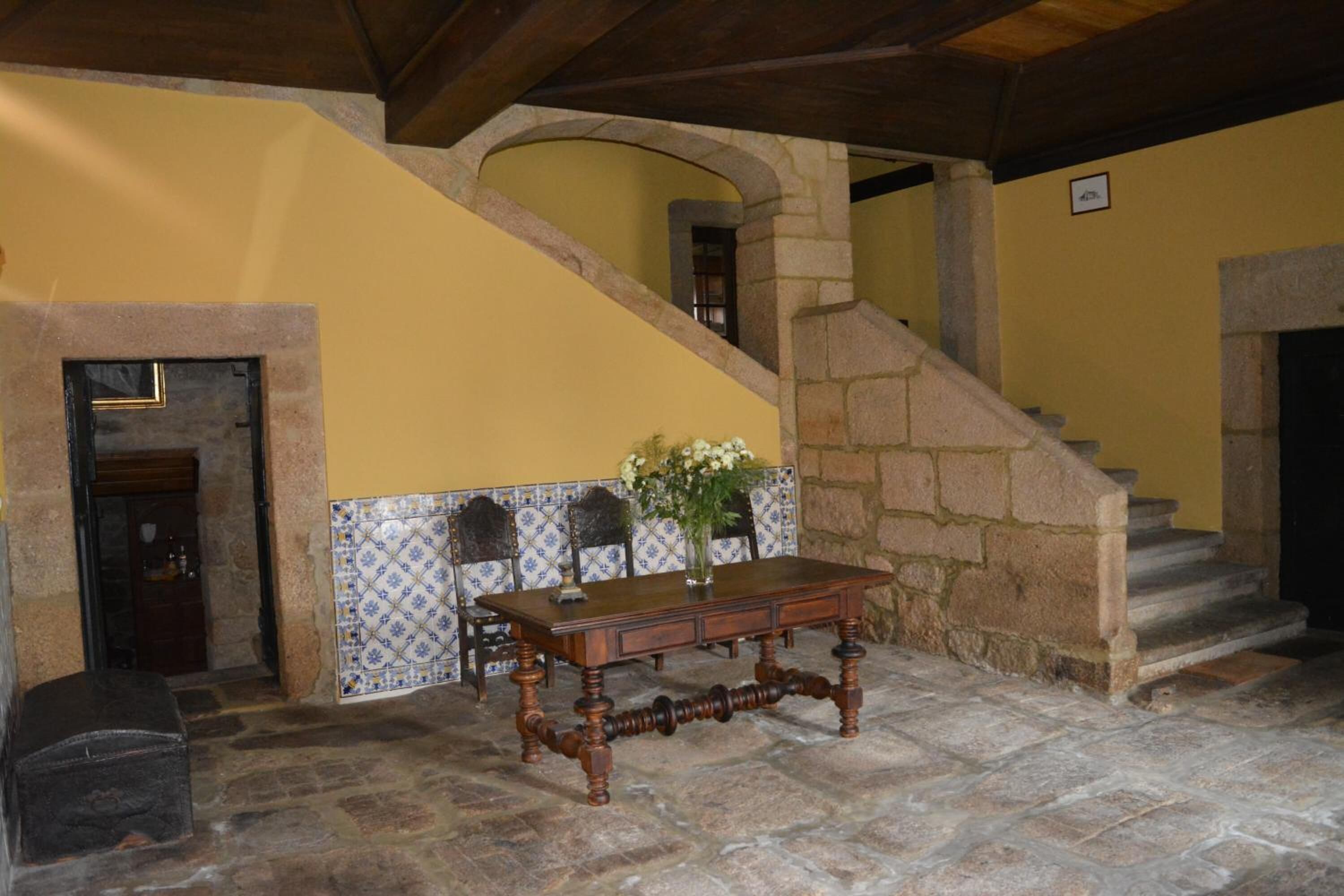 Um quarto com piso de pedra, uma mesa de madeira, um vaso de flores e parede com azulejos.