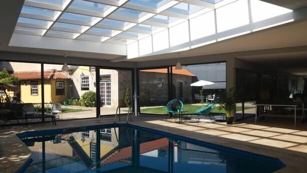 A swimming pool with a glass ceiling, large windows, and a view of a house with a red roof.