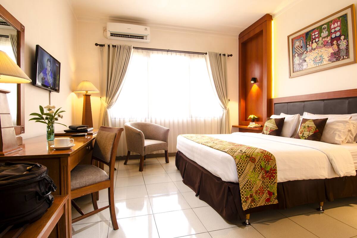 A hotel room with a bed, a desk with a chair, a TV, and a window with curtains.