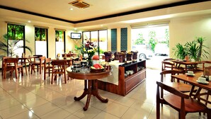A hotel dining area with wooden tables and chairs, a central round table with a flower arrangement, and a buffet counter with food items. - Griya Sentana Hotel Jogjakarta (Yogyakarta)