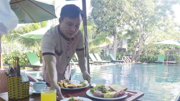 A dining table with various dishes, a glass of juice, and a waiter serving food. - Hoi An Osaka Riverside Villa & Spa (Da Nang)