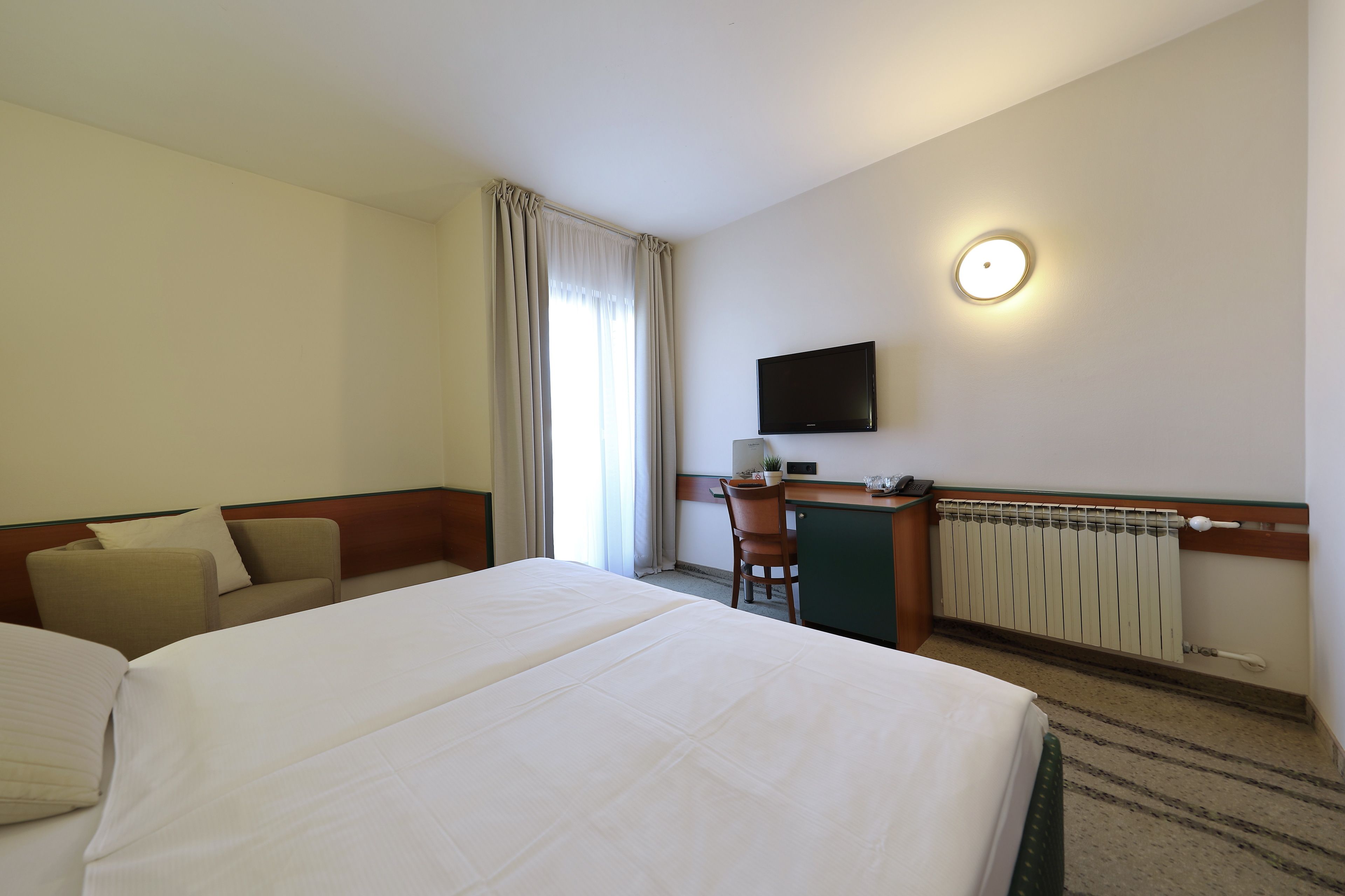 A hotel room with a large bed, a desk, a chair, a television, and a radiator.