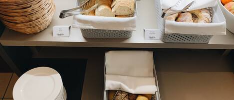Un montaje de buffet con pan, croissants y productos de repostería en cestas, junto con una pila de platos blancos.